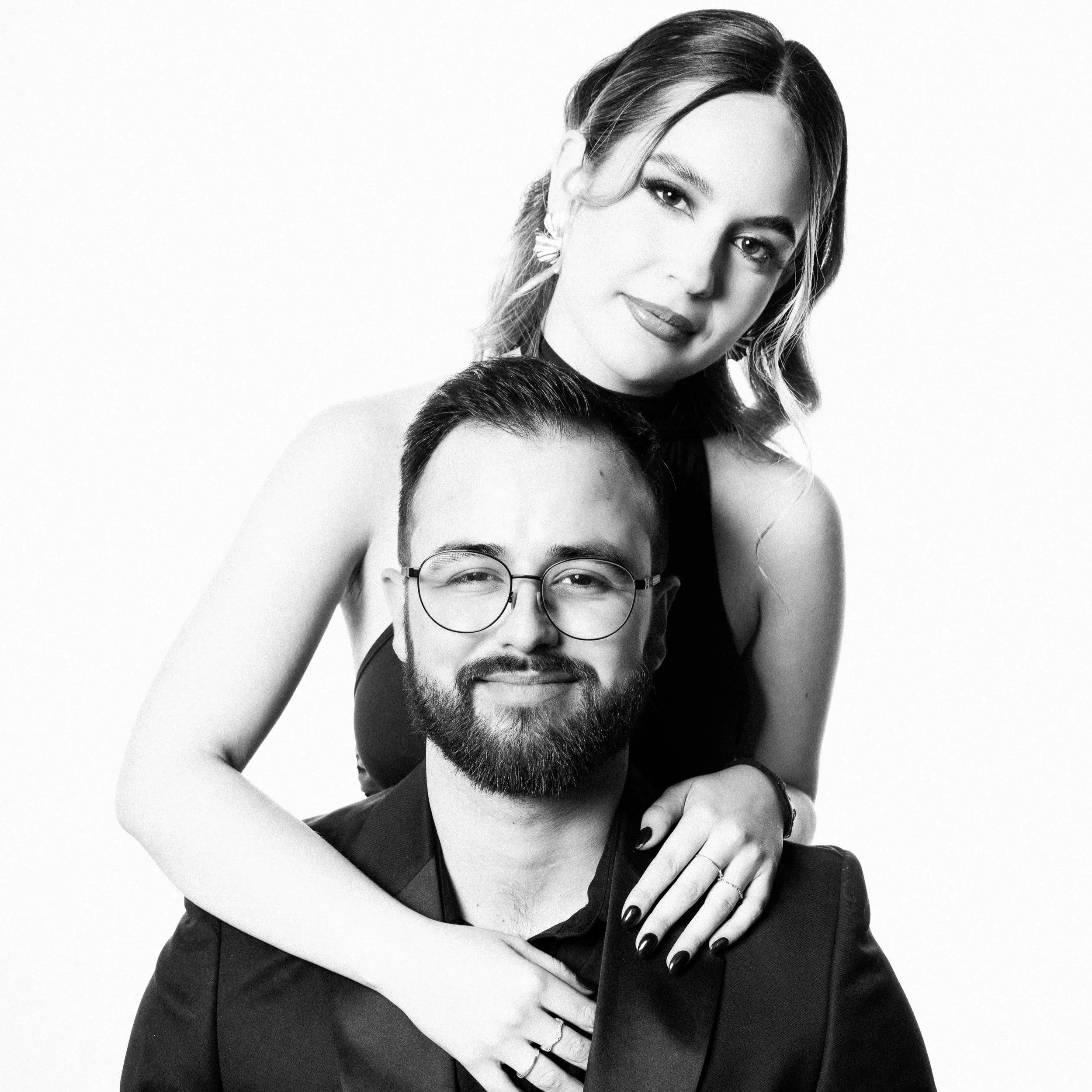 Black-and-white photograph of a woman with dark hair, styled in soft waves, and a man with glasses and a beard, smiling with their faces close together. The woman has makeup and is wearing earrings.