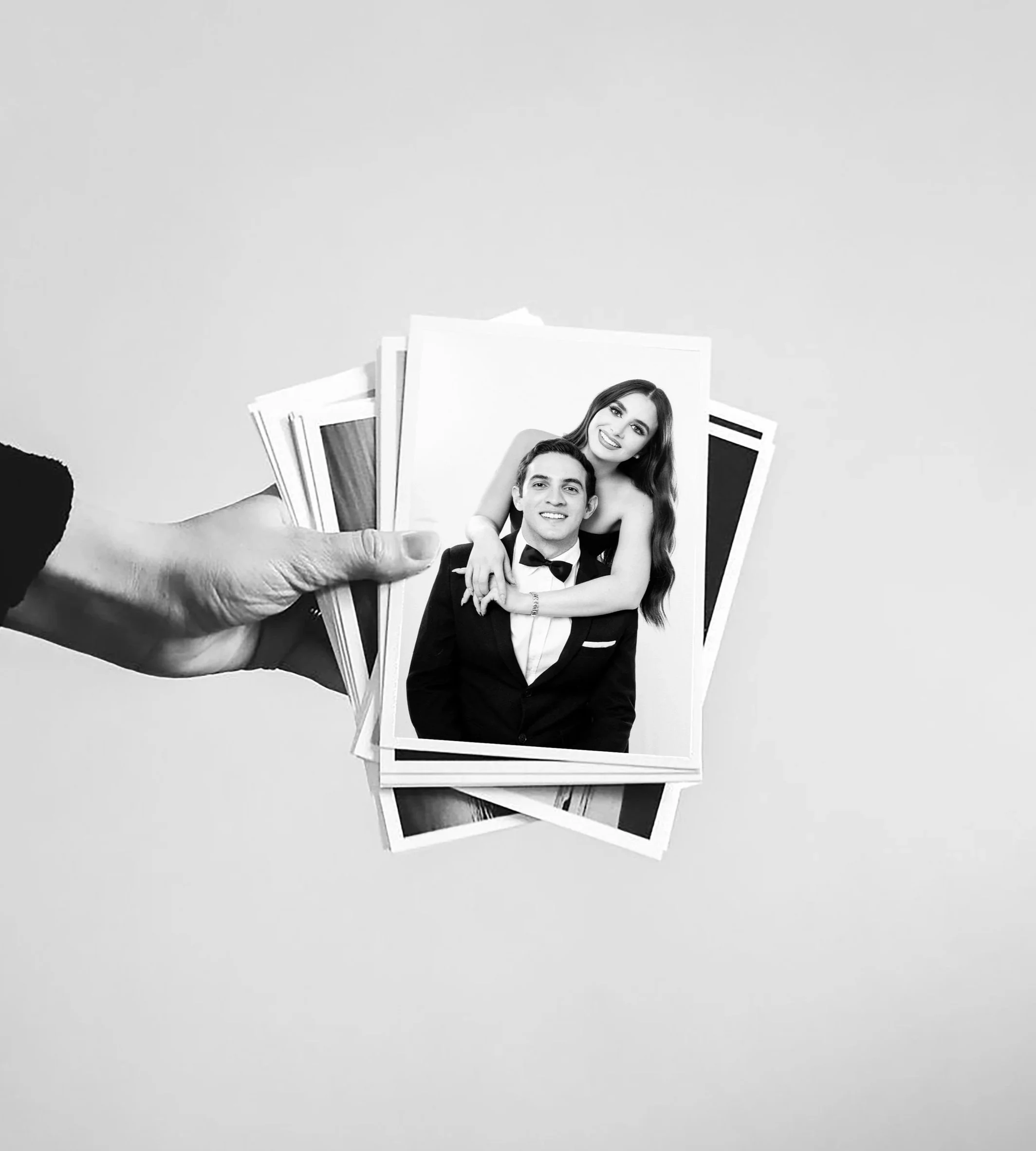 Black and white photo of a hand holding several printed photographs of a smiling couple and a woman, spread out against a plain background.