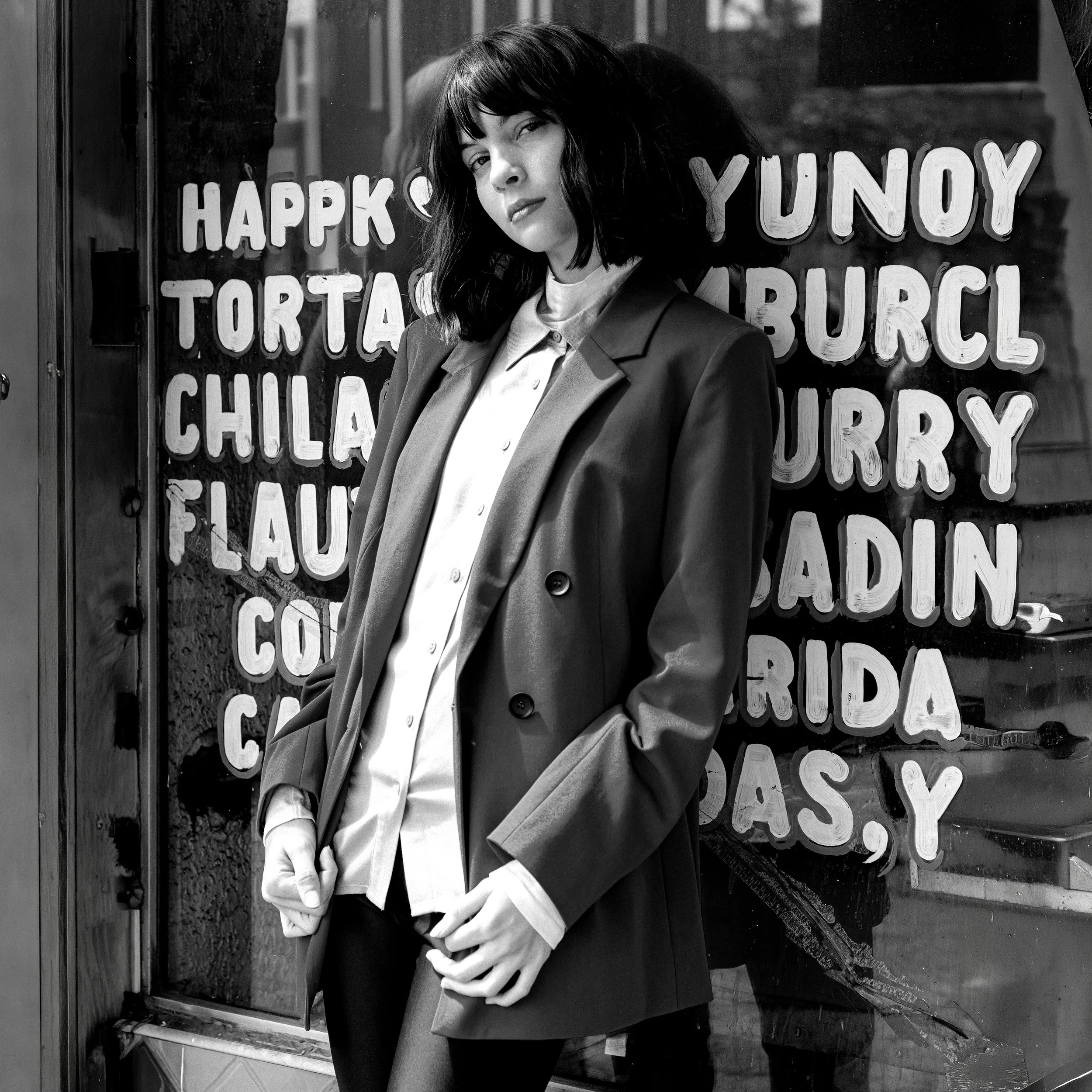 A woman with black hair and bangs leaning her head against a glass window with a sign behind her that reads 'HAPPY YOUTH' and other words. She is wearing a suit jacket over a shirt and is standing outside.