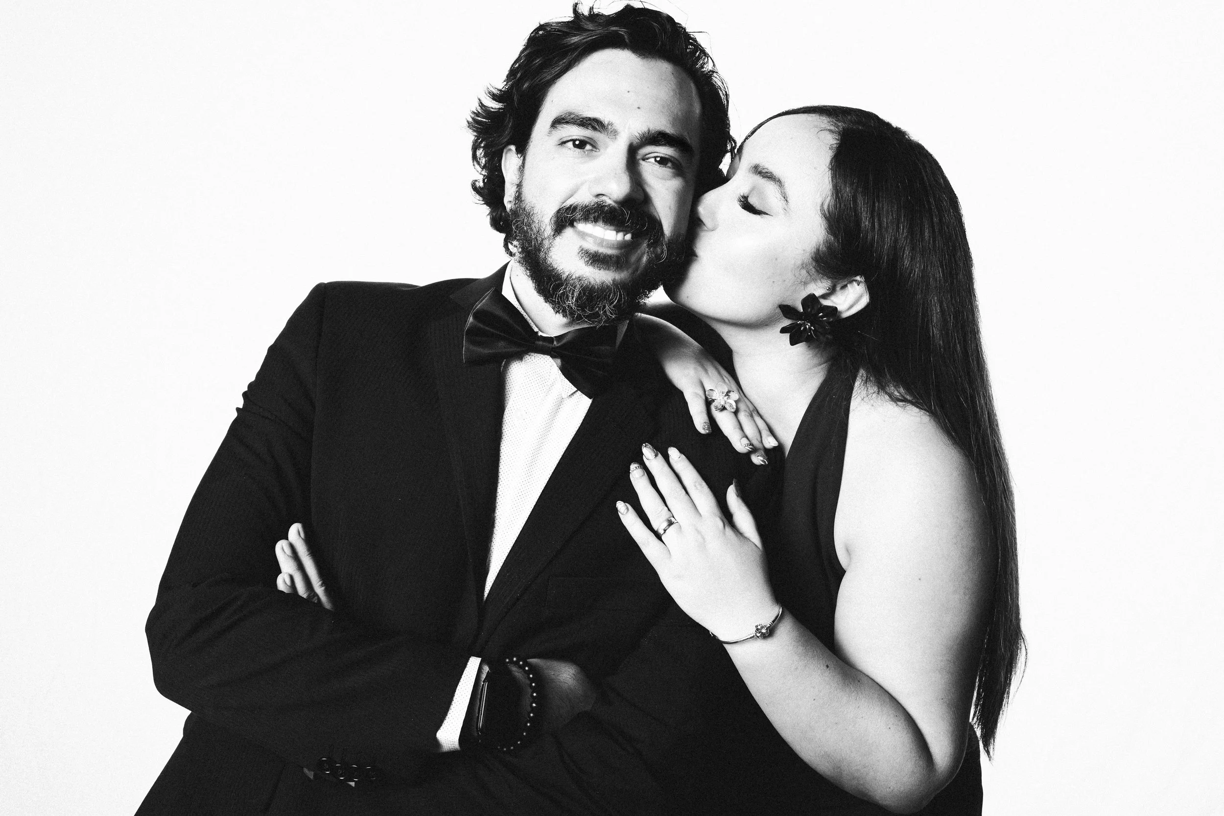 A black-and-white photo of a man in a tuxedo and a woman in a dress, with the woman kissing the man's cheek. The man has a beard and mustache, and the woman has long dark hair and is wearing earrings and rings.