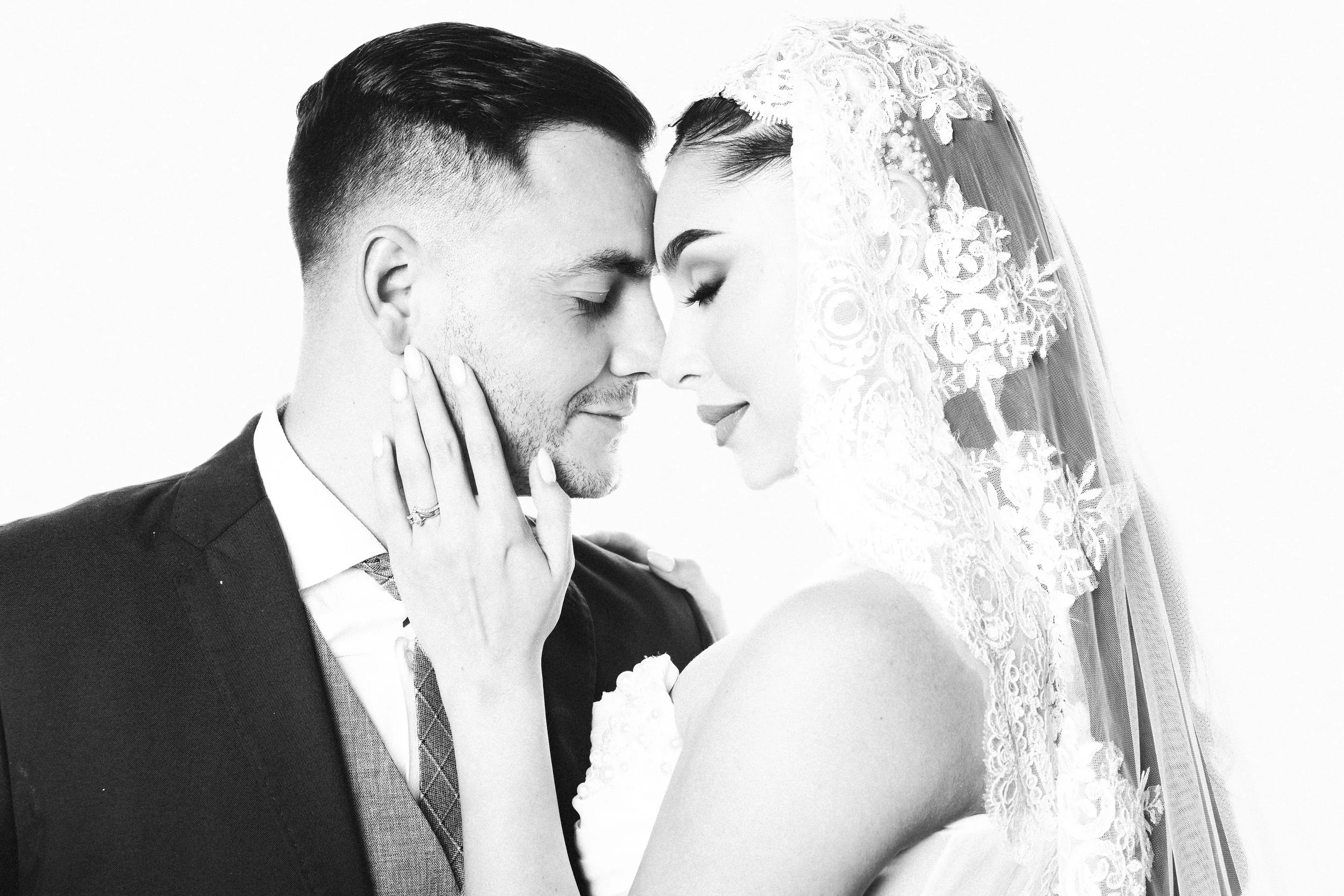 Black and white photo of a bride and groom close together, touching foreheads, with eyes closed, a wedding ring visible on the bride's finger, and the bride wearing a lace veil.