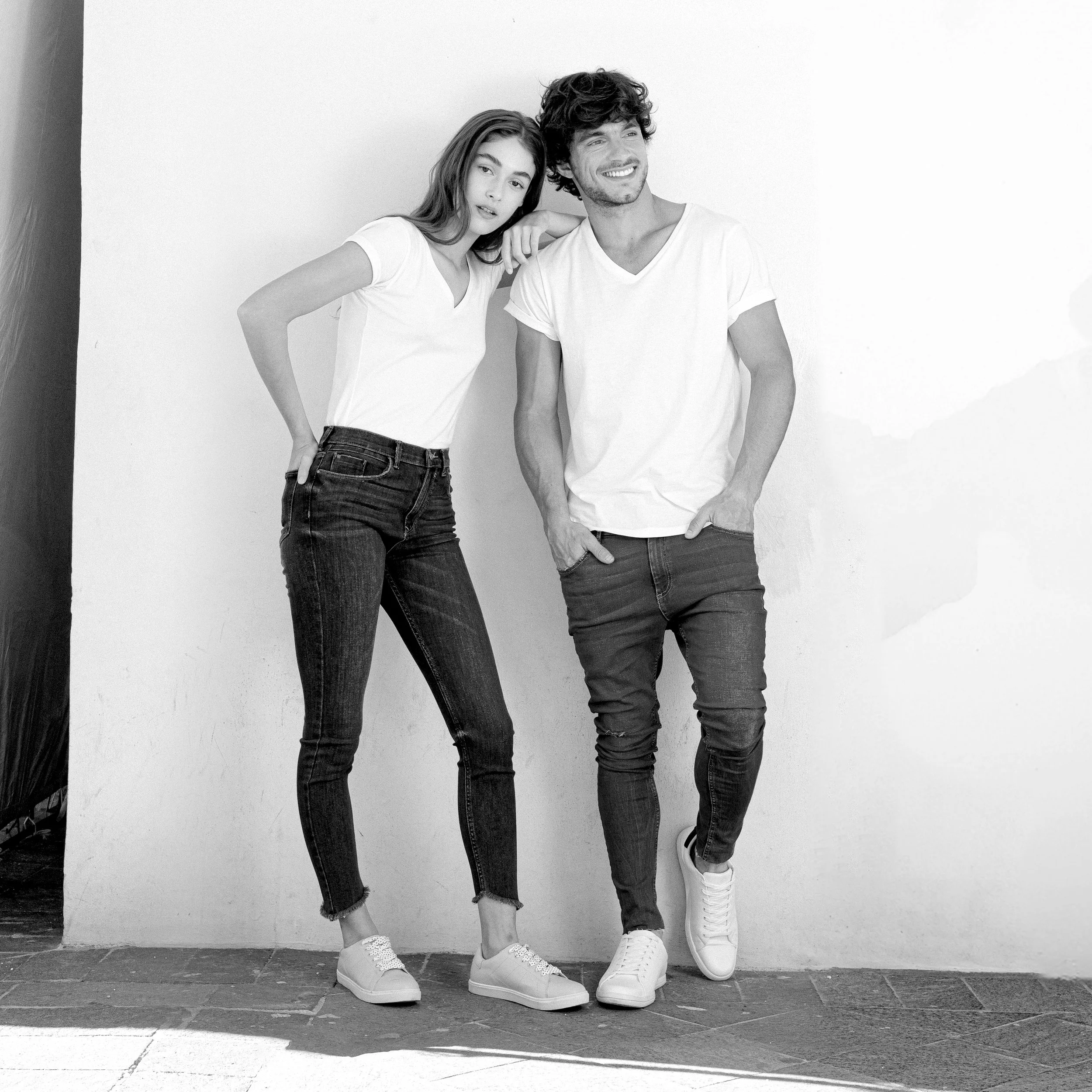 A young woman and young man standing close to each other against a plain wall, wearing casual white t-shirts, jeans, and sneakers, smiling and posing for a photo.