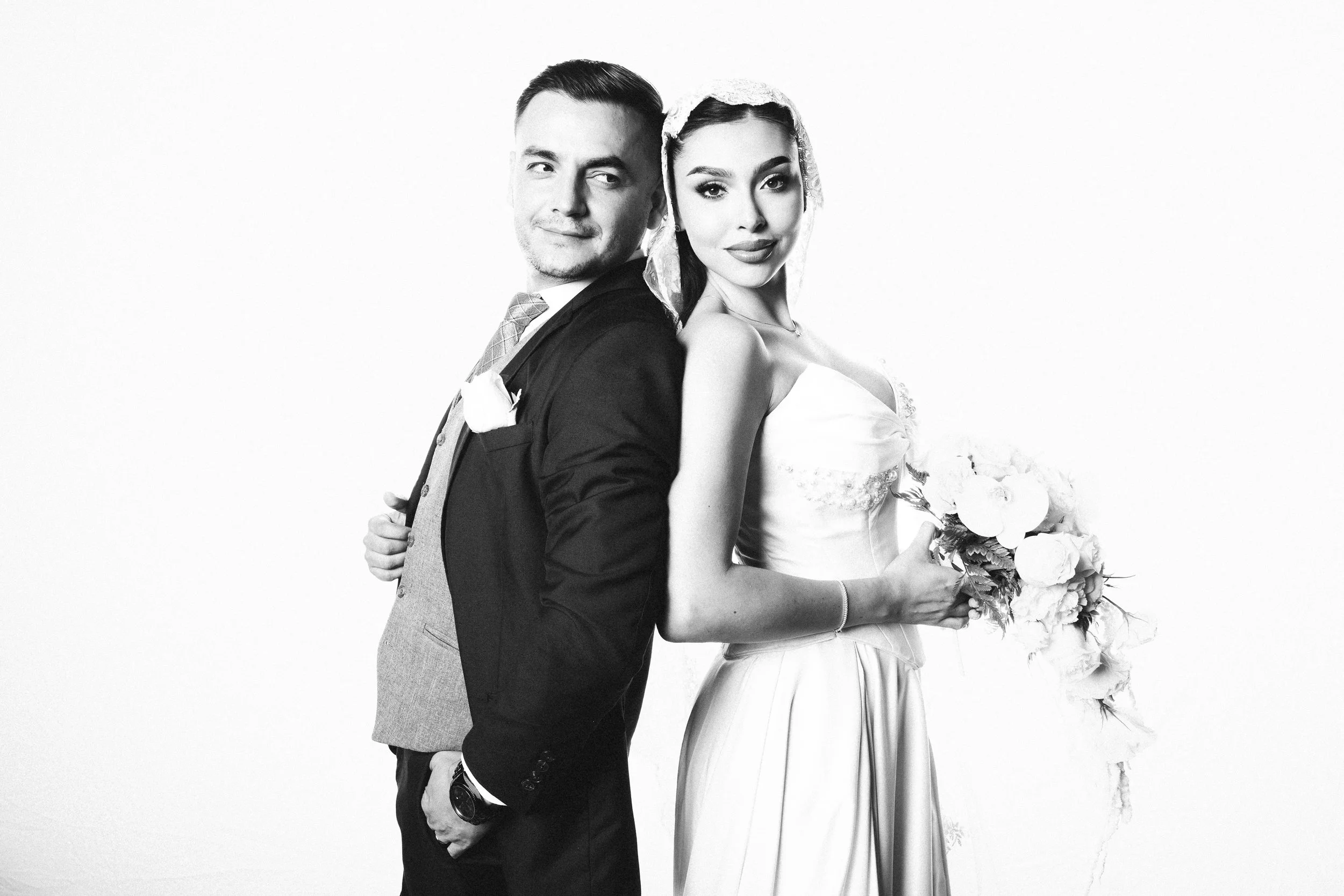 A black-and-white photo of a couple on their wedding day. The groom is dressed in a suit, and the bride is wearing a wedding dress and holding a bouquet of flowers.