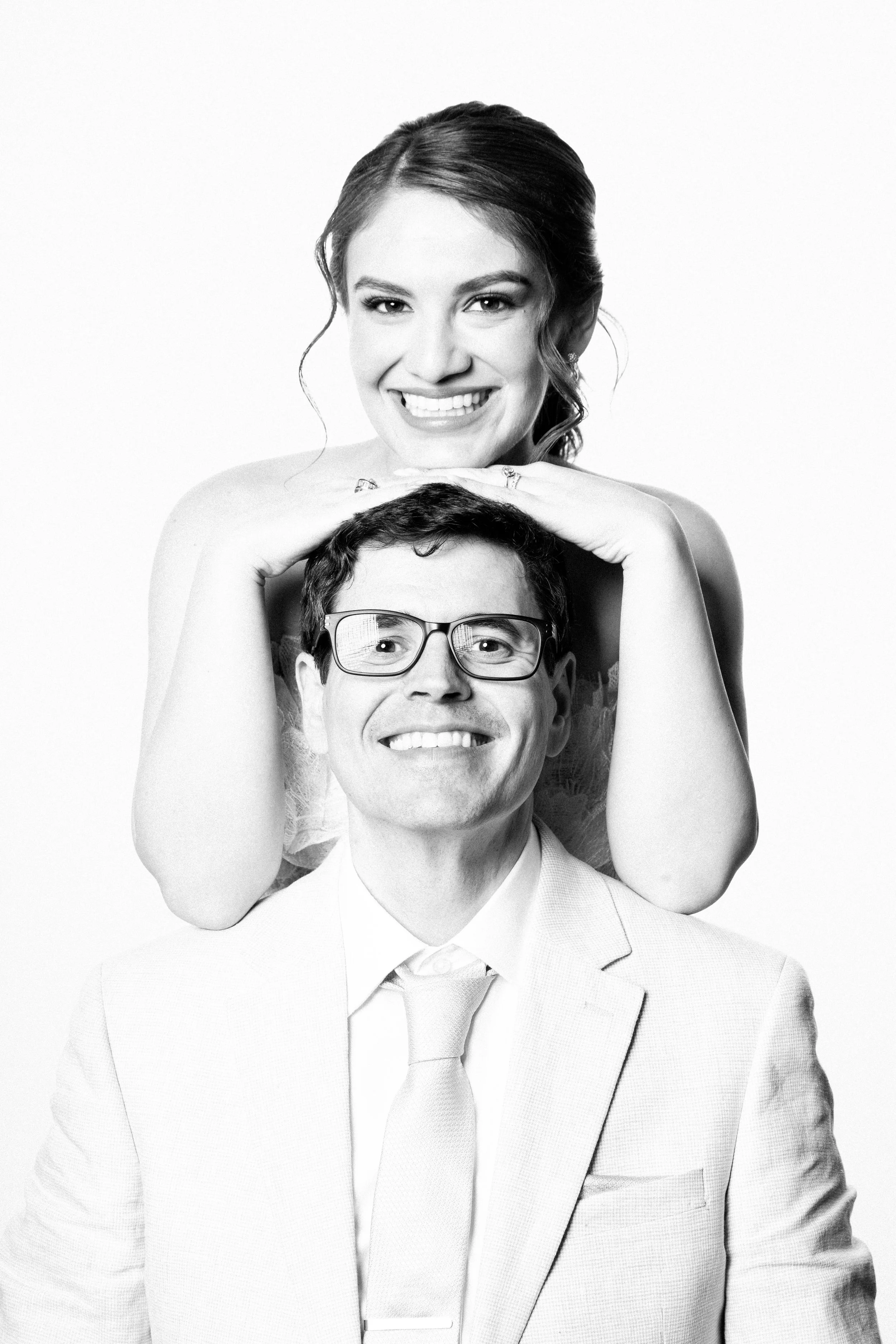 Black-and-white photo of a woman smiling, with her arms resting on the shoulders of a man in a suit and glasses, who is smiling and looking at the camera.