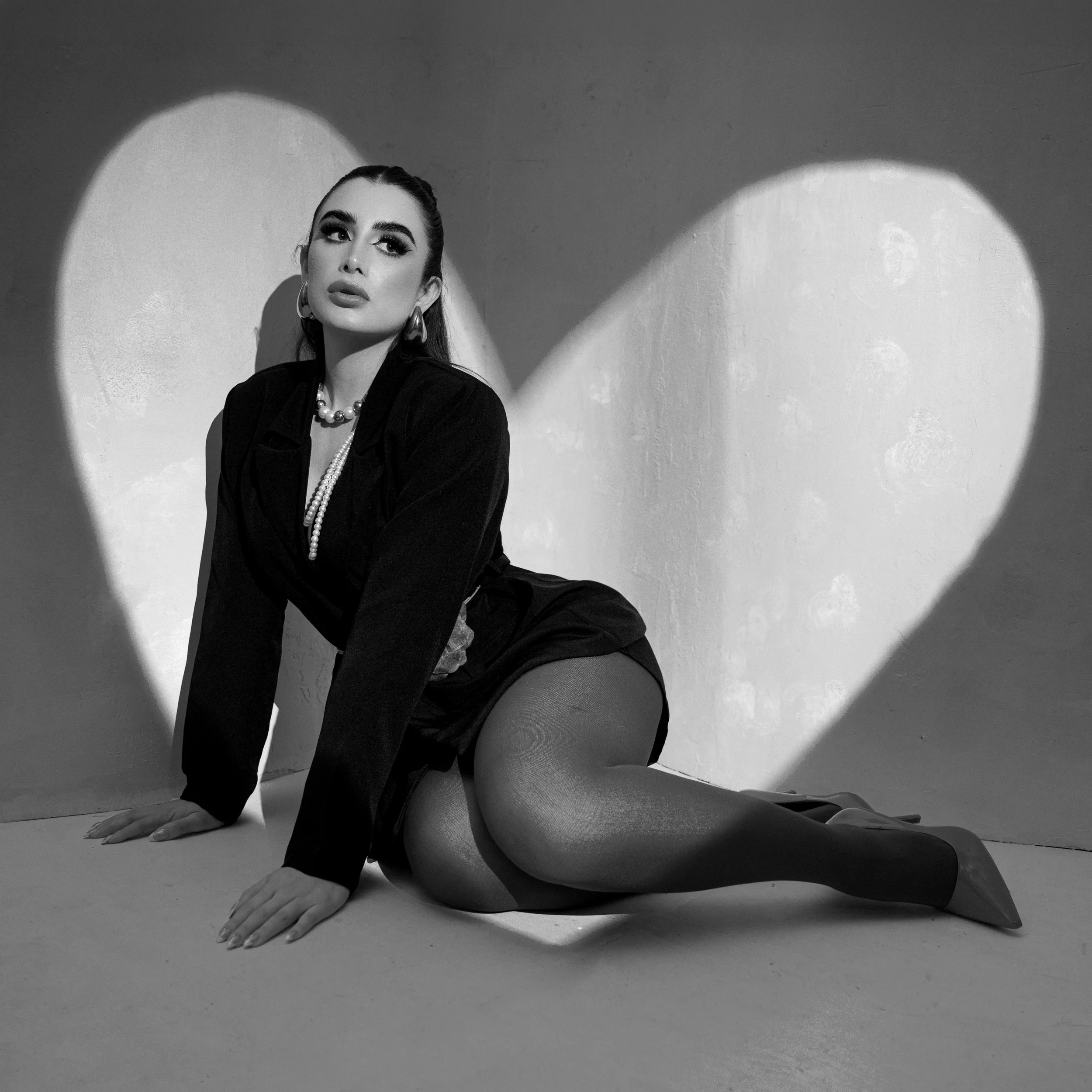 A black-and-white photo of a woman with dark hair in a bun, wearing a dark jacket, pearl necklace, and hoop earrings, posing with her hands on the ground as she sits on her knees against a plain wall with circular light patches.