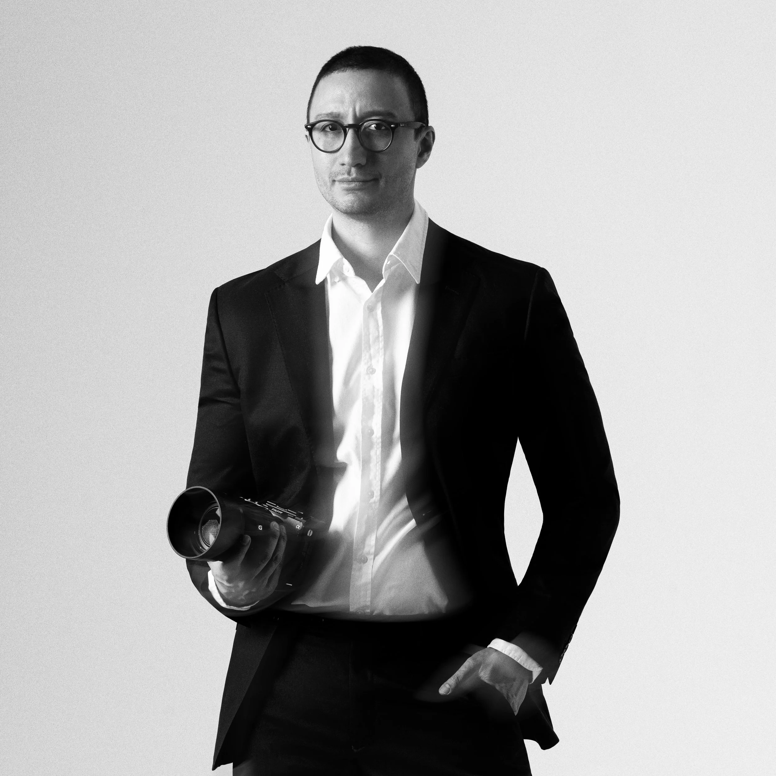 Black and white portrait of a man wearing glasses, a white shirt, and a dark blazer, holding a camera, against a plain background.