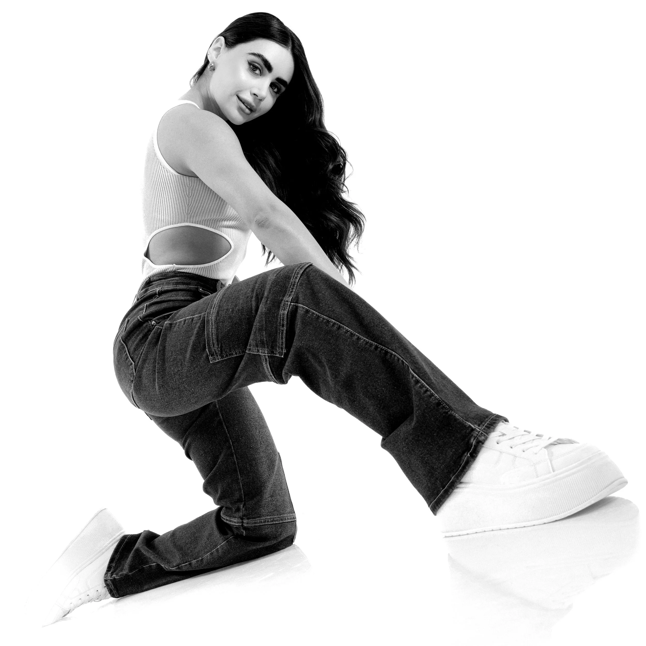 A woman with long dark hair, wearing a sleeveless top and jeans, sitting on the ground with her legs crossed, smiling at the camera.