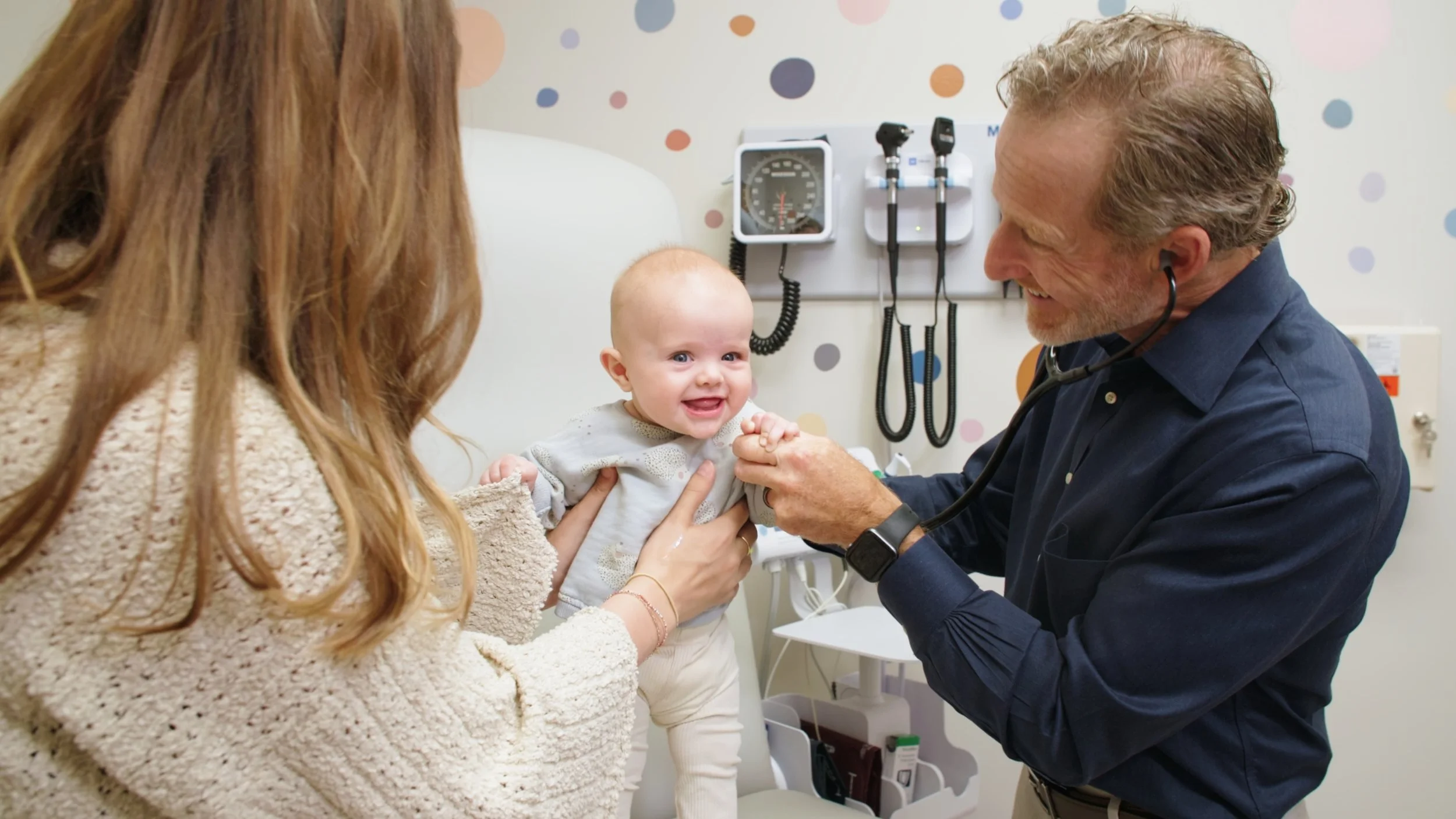 A young baby with a bald head smiling with the help of a woman, while a male doctor with a stethoscope examines the baby in a medical examination room.
