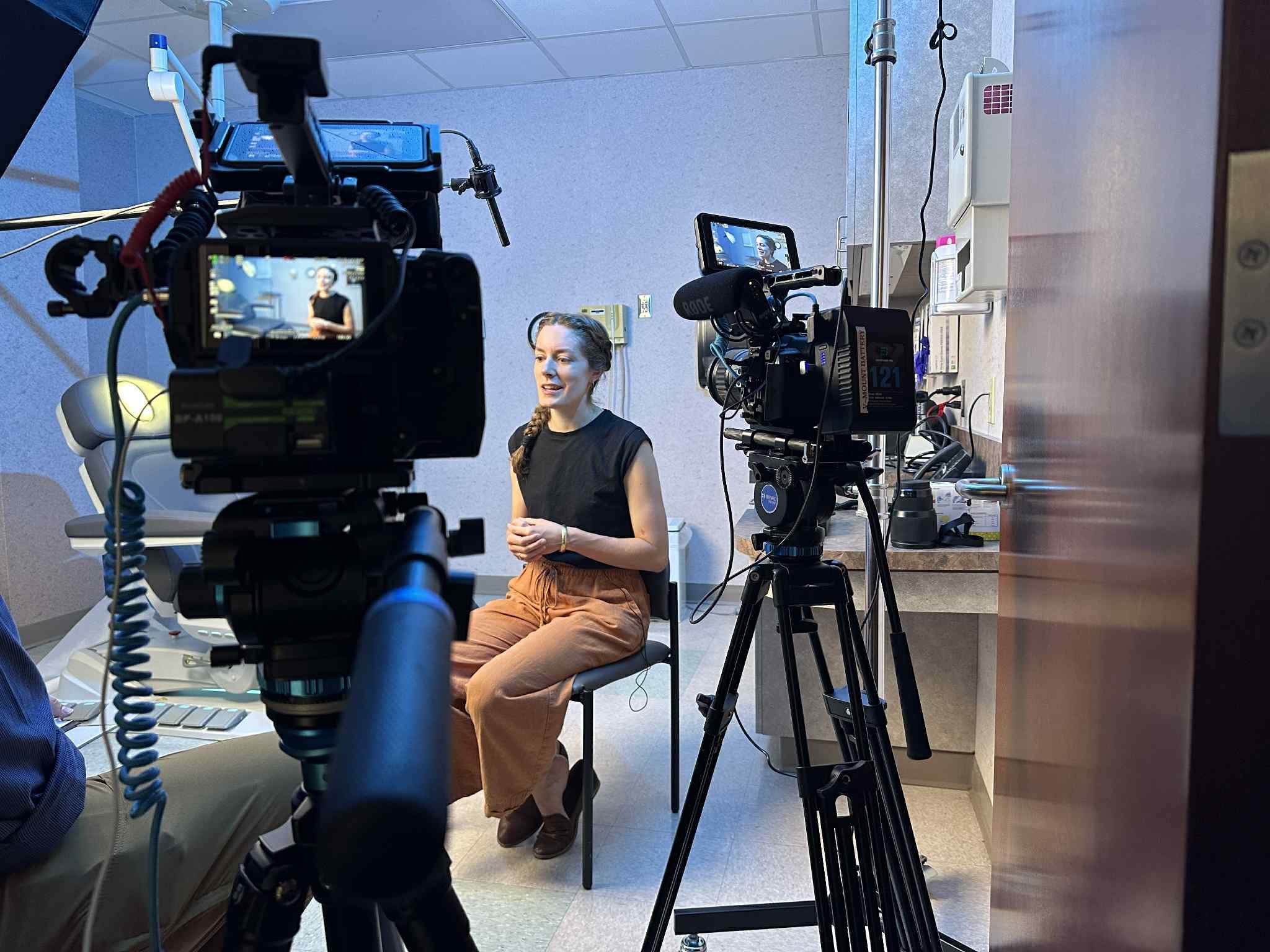 A woman sitting in a medical setting, being filmed by multiple professional cameras. She has braided hair and is wearing an orange skirt and a black top.