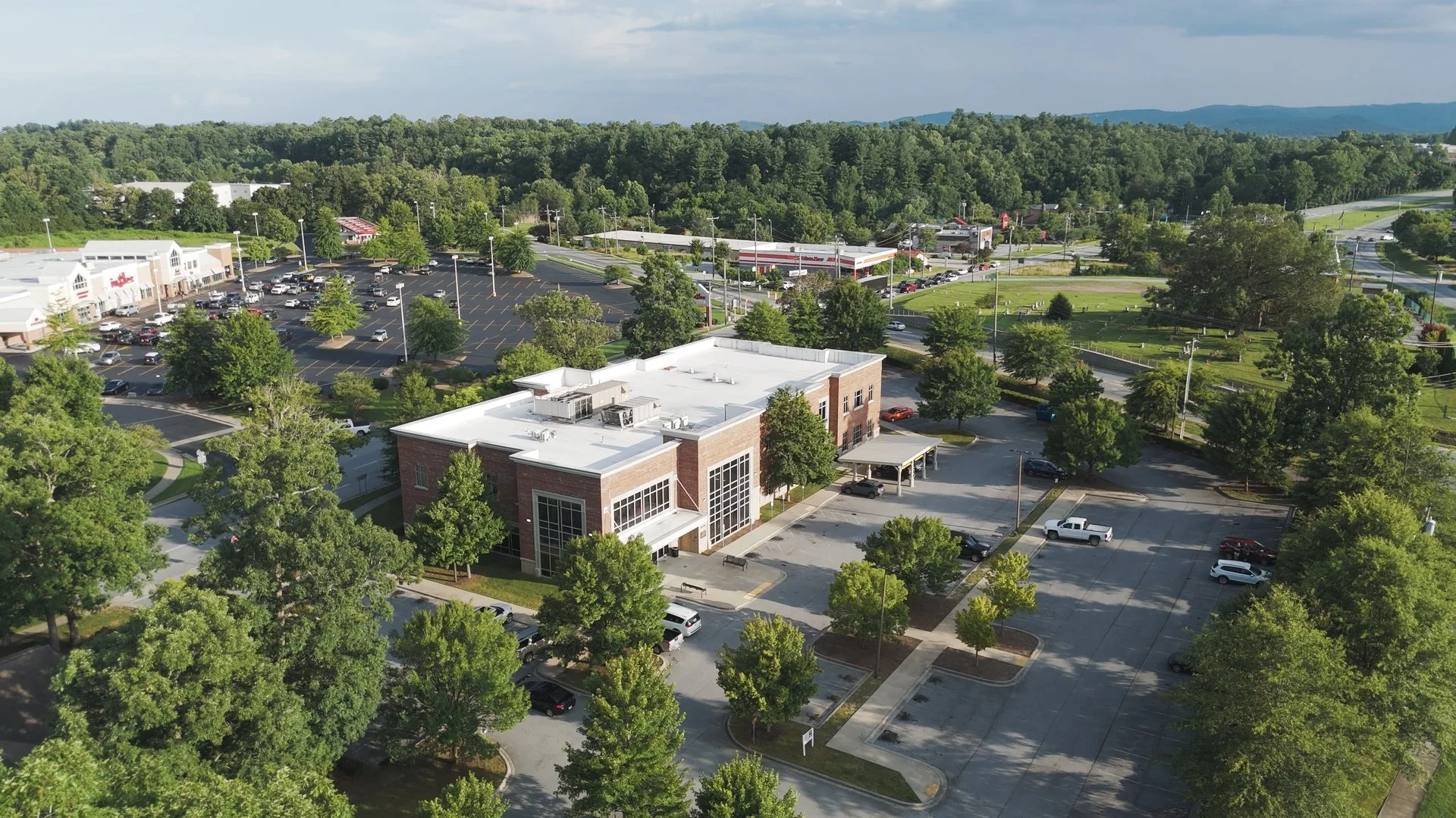 Aerial view of a commercial shopping center with storefronts, parking lots, and surrounding trees, with a wooded area and hills in the background.