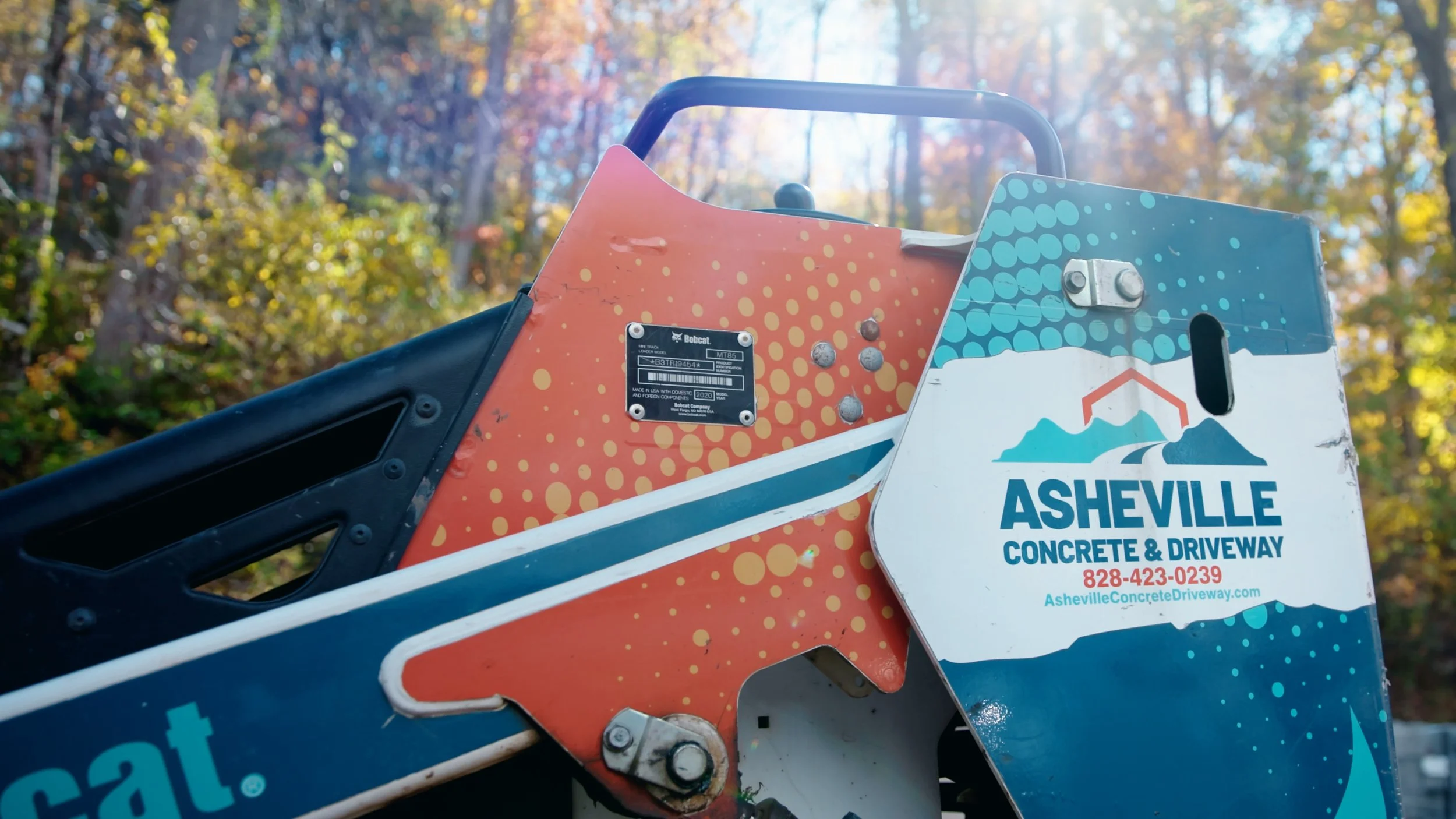 Close-up of a construction sign for Asheville Concrete & Driveway showing contact information, attached to a piece of machinery, with a background of blurred trees and sunlight filtering through leaves.
