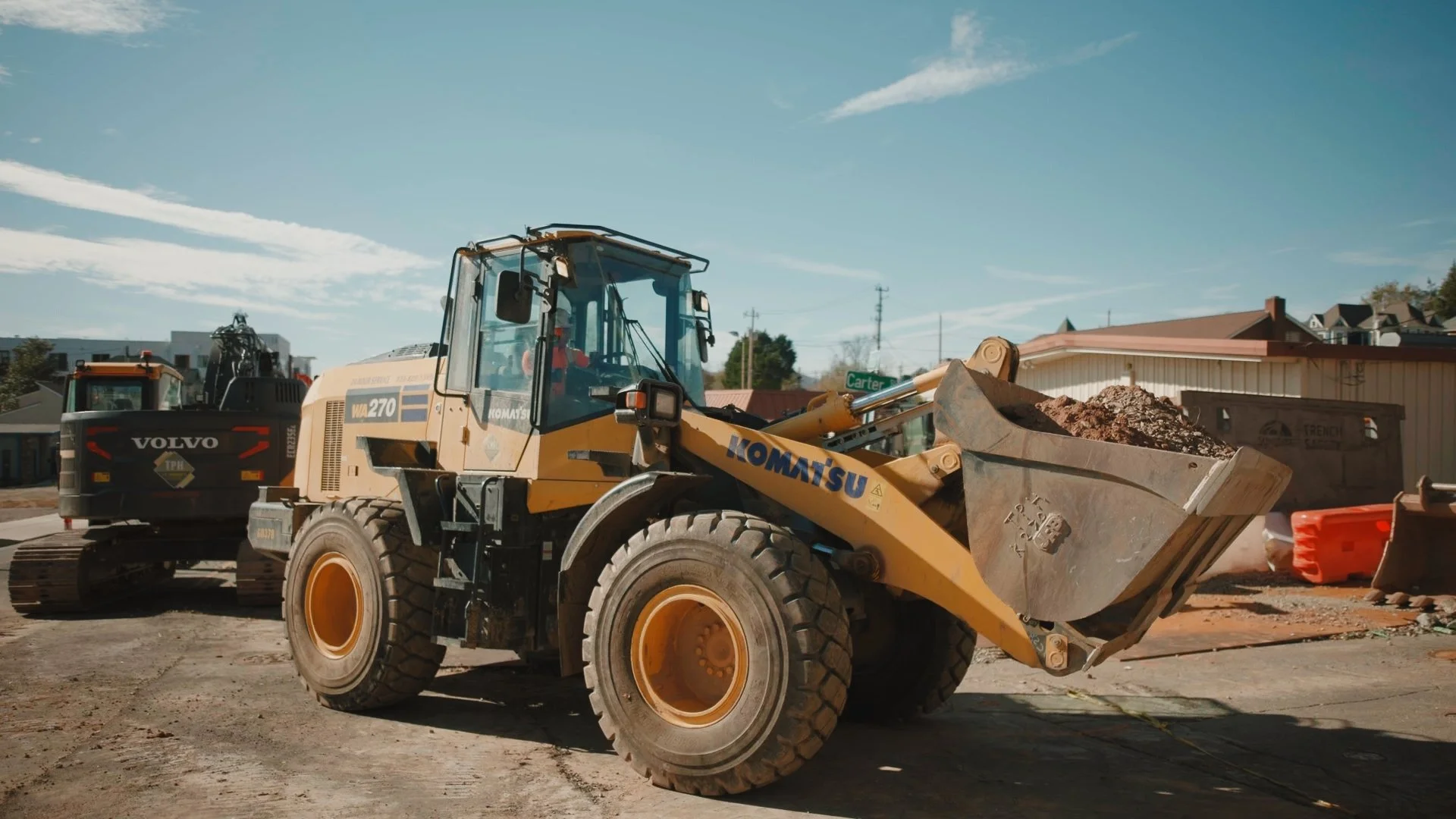 A construction site with a Komatsu front loader carrying dirt, a Volvo excavator in the background, and construction materials and debris around, under a blue sky.