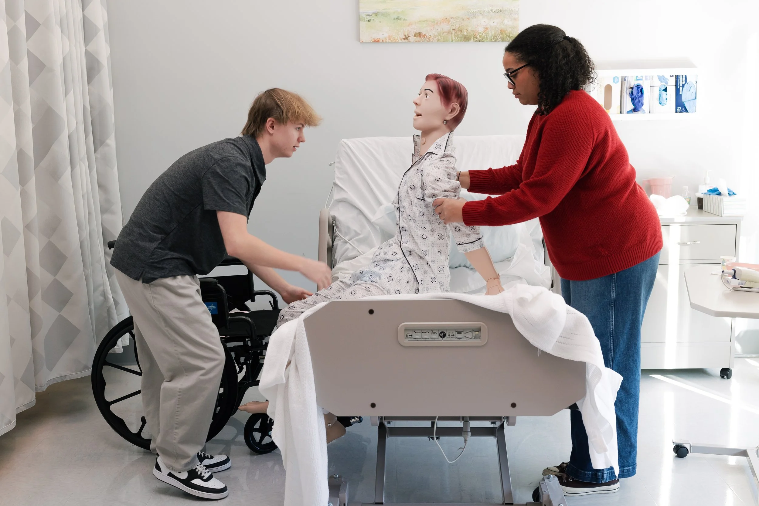 A young man in a wheelchair leaning toward a hospital bed, talking to a female healthcare worker with glasses and curly hair, who is holding a medical mannequin. A nurse or patient care assistant stands beside the bed holding the mannequin's arm, while a patient with red hair lies in the bed. The setting is a hospital room with medical supplies and artwork on the wall.