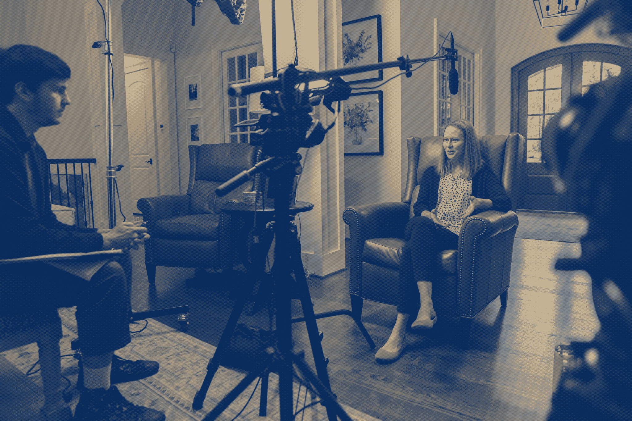 People on a film set with equipment and a camera, inside a room with large windows.