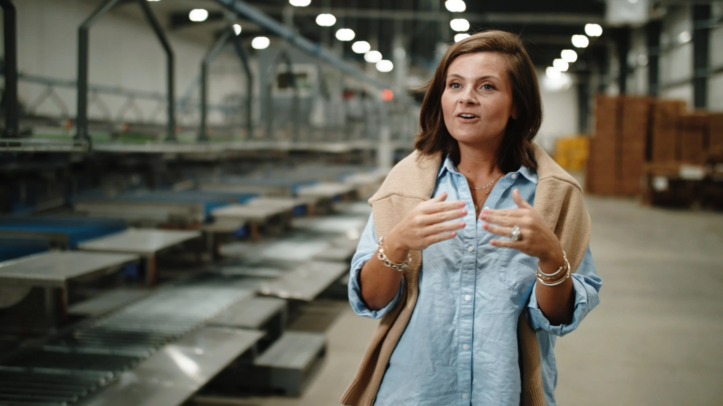 A woman speaking or explaining something inside an industrial or warehouse setting with metal shelves and equipment in the background.