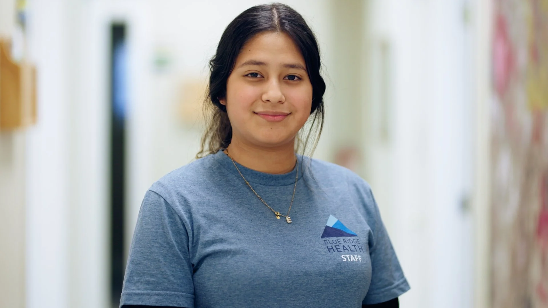 A young woman with dark hair pulled back, wearing a blue t-shirt with 'Blue Ridge Health Staff' printed on it, smiling softly, standing in a hallway with a blurred background.