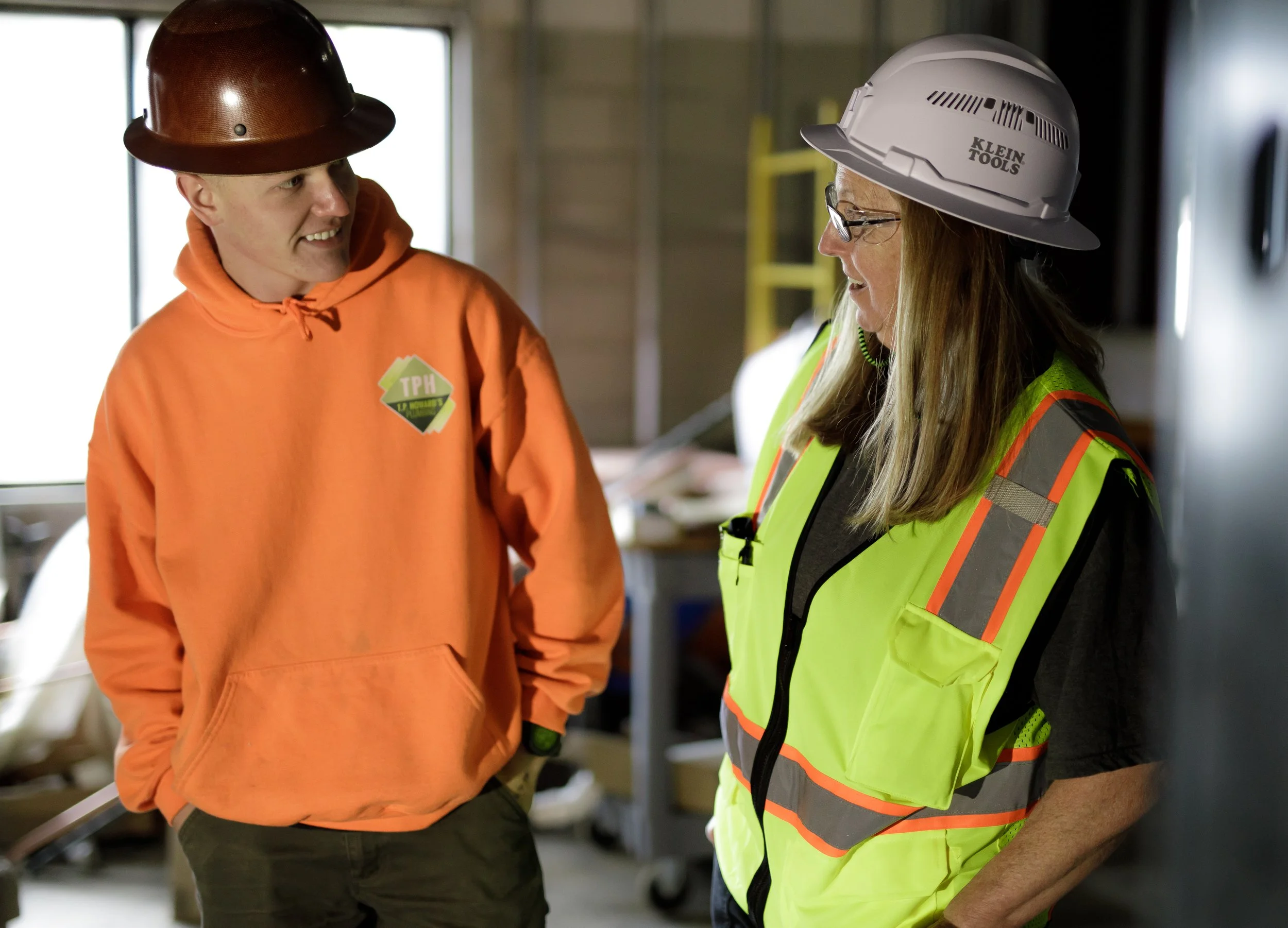 Two construction workers, a man and a woman, talking inside a building under construction. The man is wearing an orange hoodie and a brown hard hat, while the woman is wearing a high-visibility safety vest, glasses, and a white hard hat.