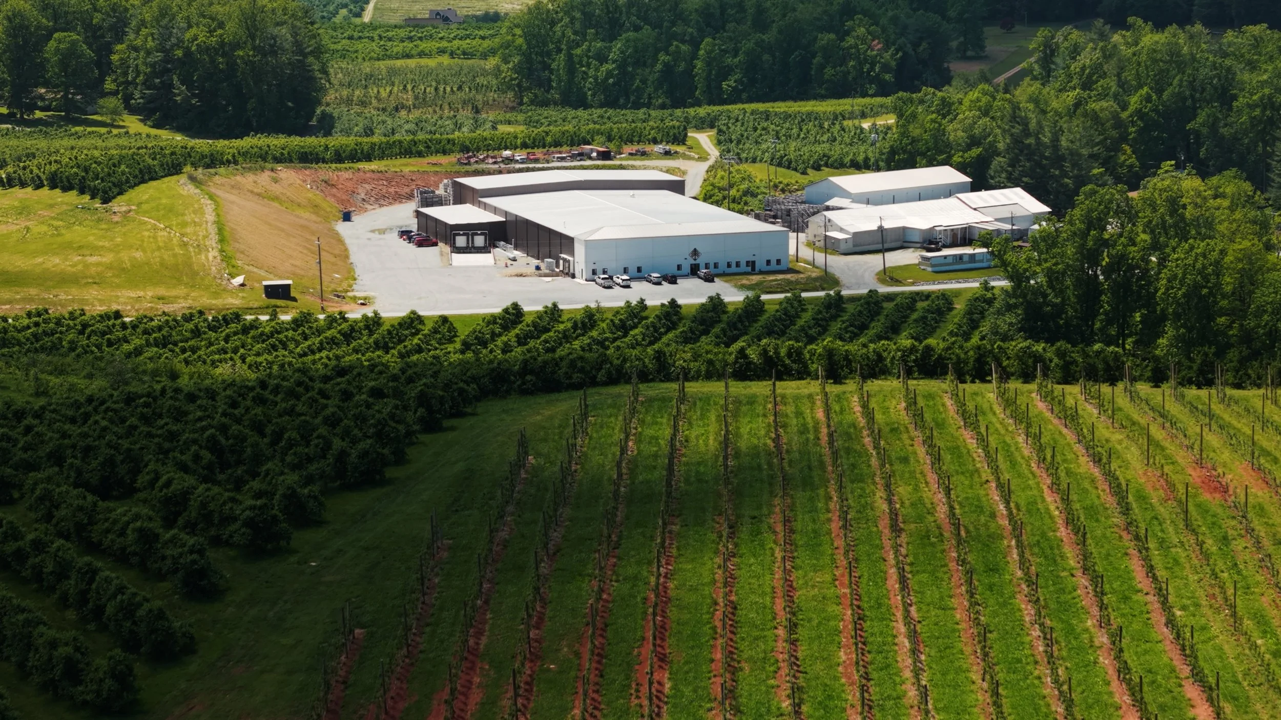 An aerial view of a large white industrial building surrounded by green farmland, vineyards, and dense trees.