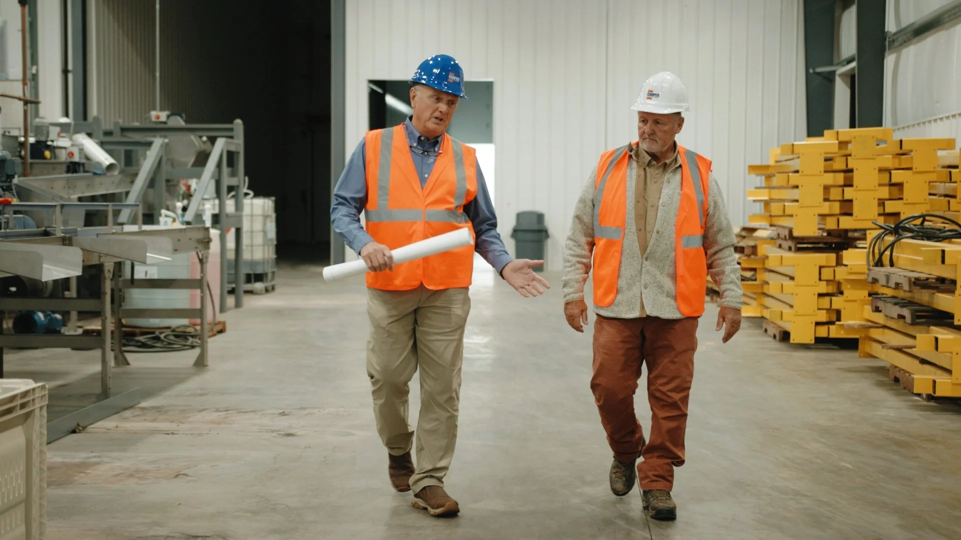 Two men wearing orange safety vests and hard hats walking inside a warehouse or factory. One man is holding blueprints and appears to be explaining something to the other.