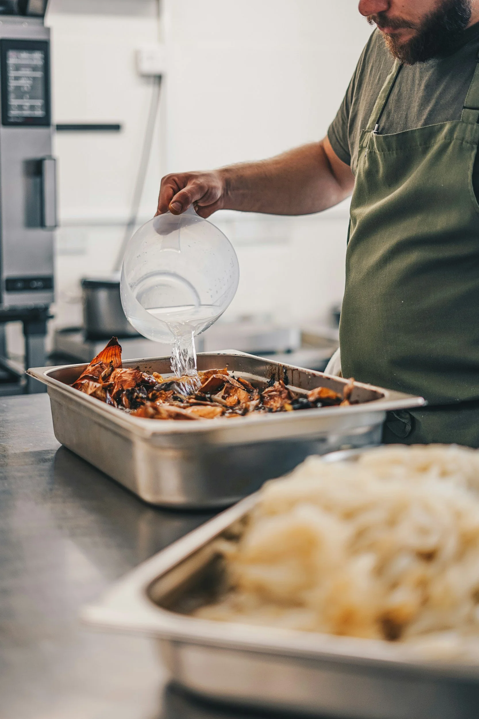 A man with a beard wearing an apron pouring water into a baking dish filled with cooked seafood in a professional kitchen.