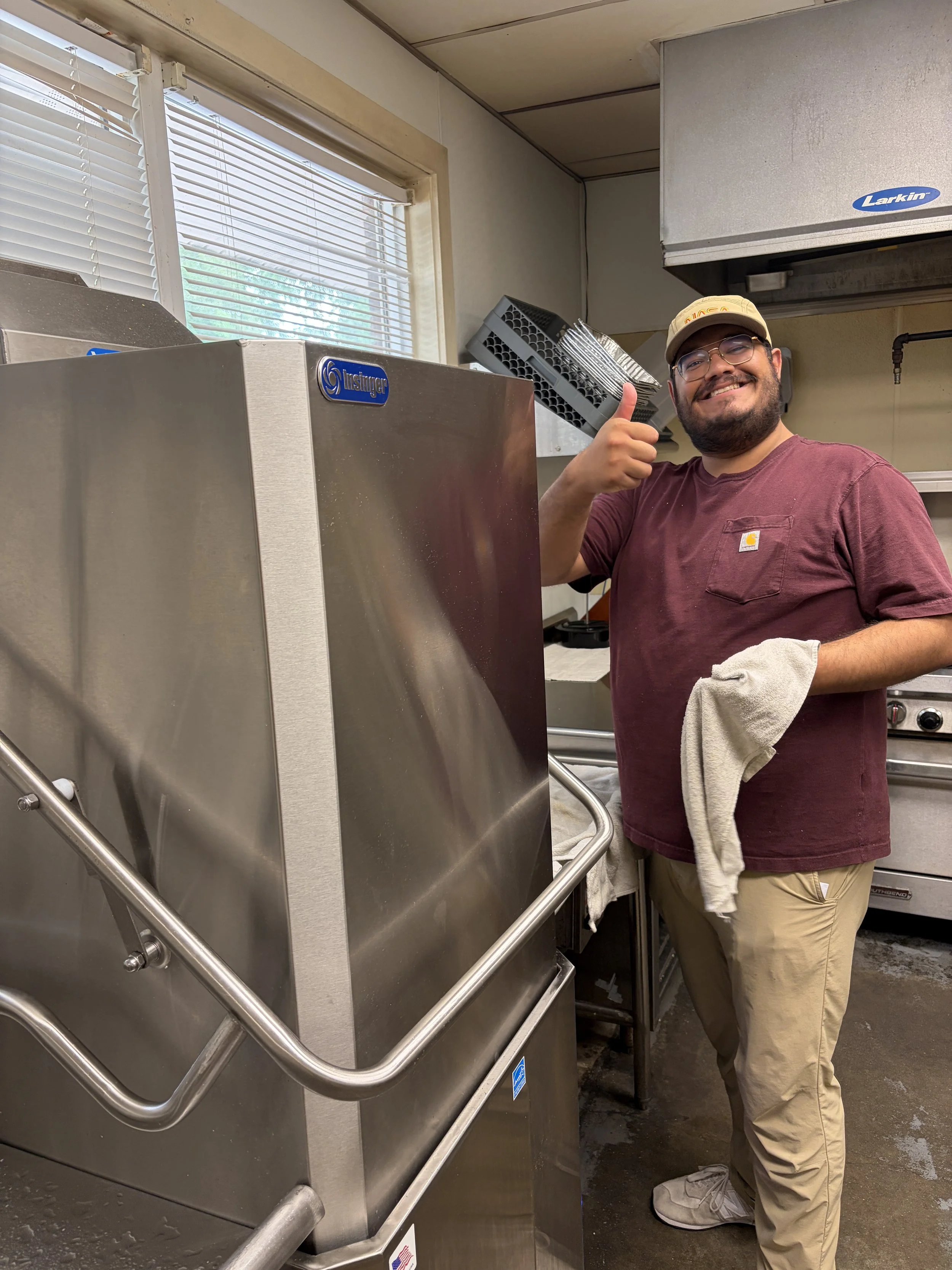 A man wearing glasses, a beige baseball cap, a maroon T-shirt, and beige pants is smiling and giving a thumbs up while standing next to a stainless steel commercial dishwasher in a kitchen.