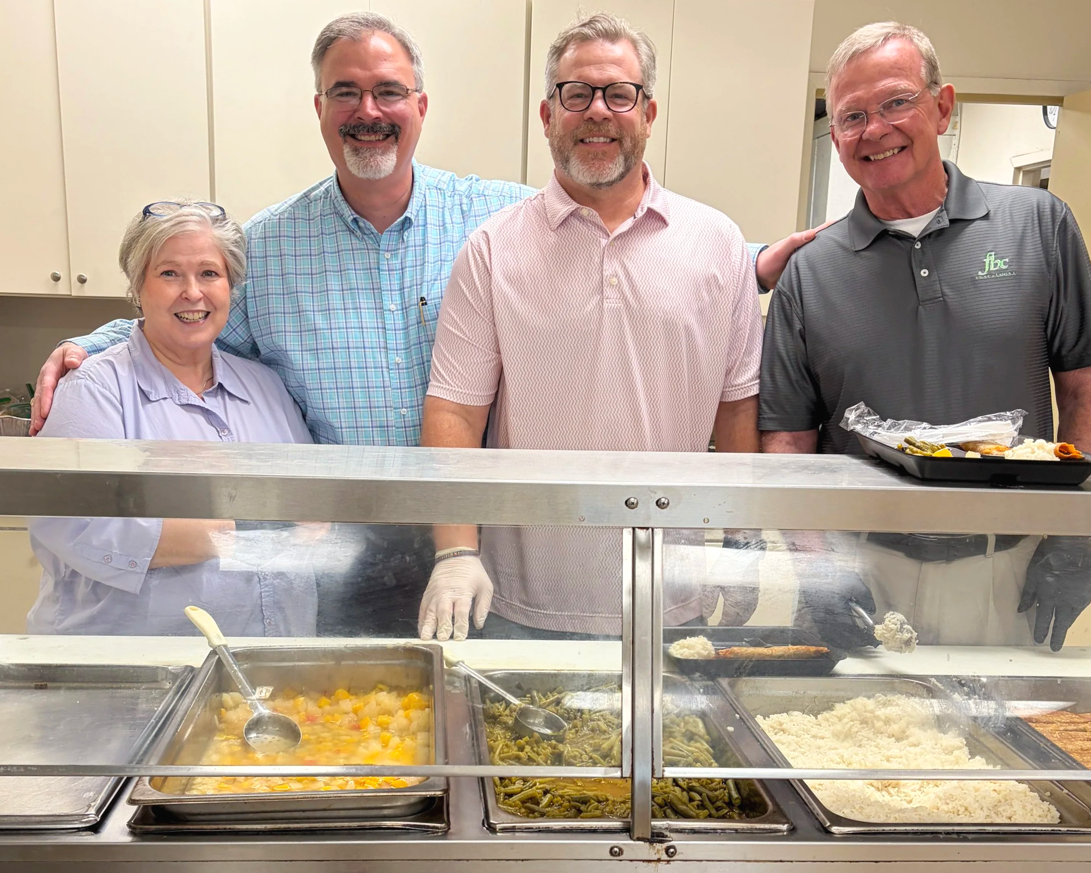 Four people, two men and a woman, smiling inside a cafeteria or food service setting. They are standing behind a serving counter with trays of food including mashed potatoes, green beans, and a vegetable medley.