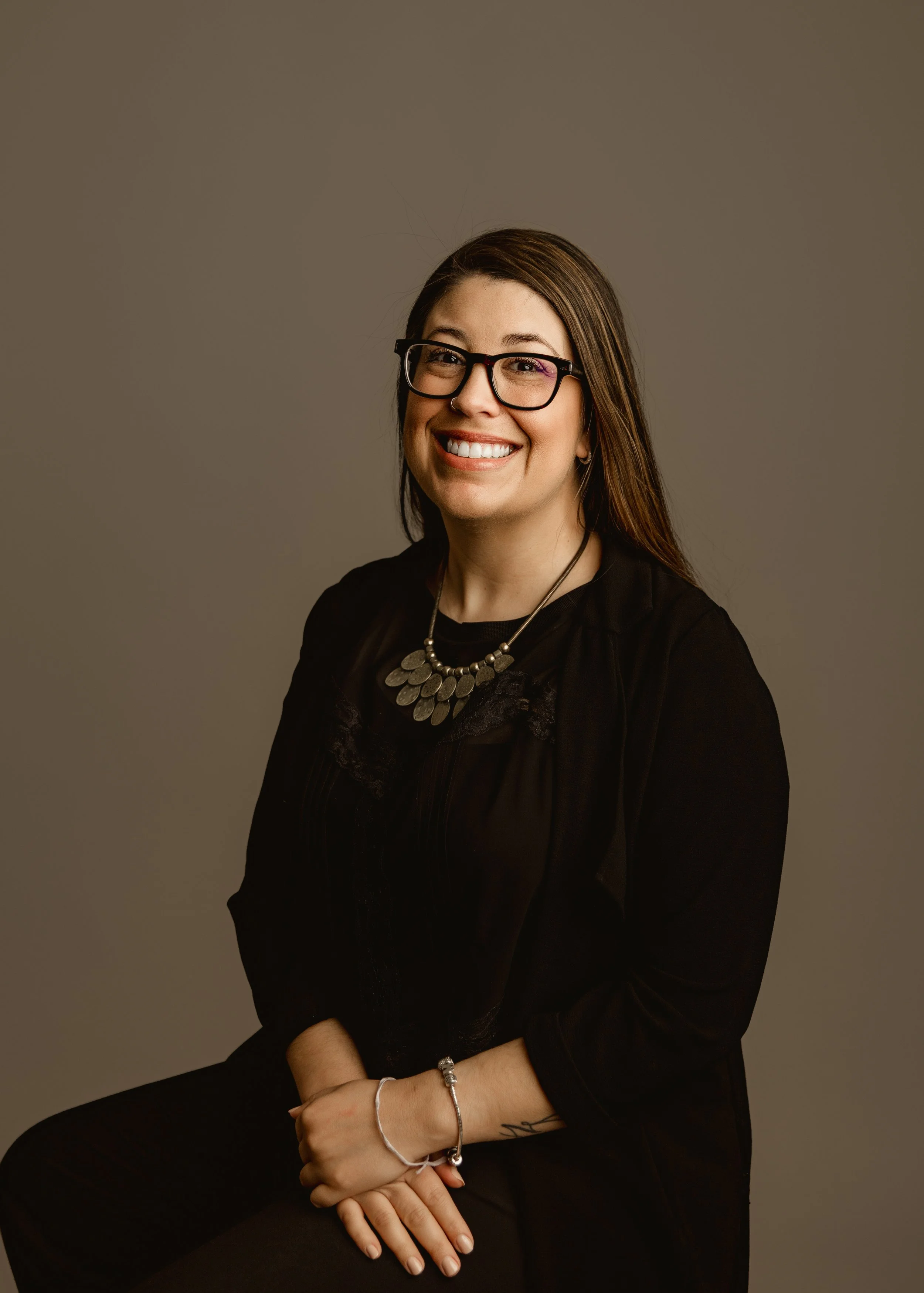 Portrait of a woman with long brown hair, glasses, and a nose ring, smiling, wearing a black top and statement necklace, against a neutral background.