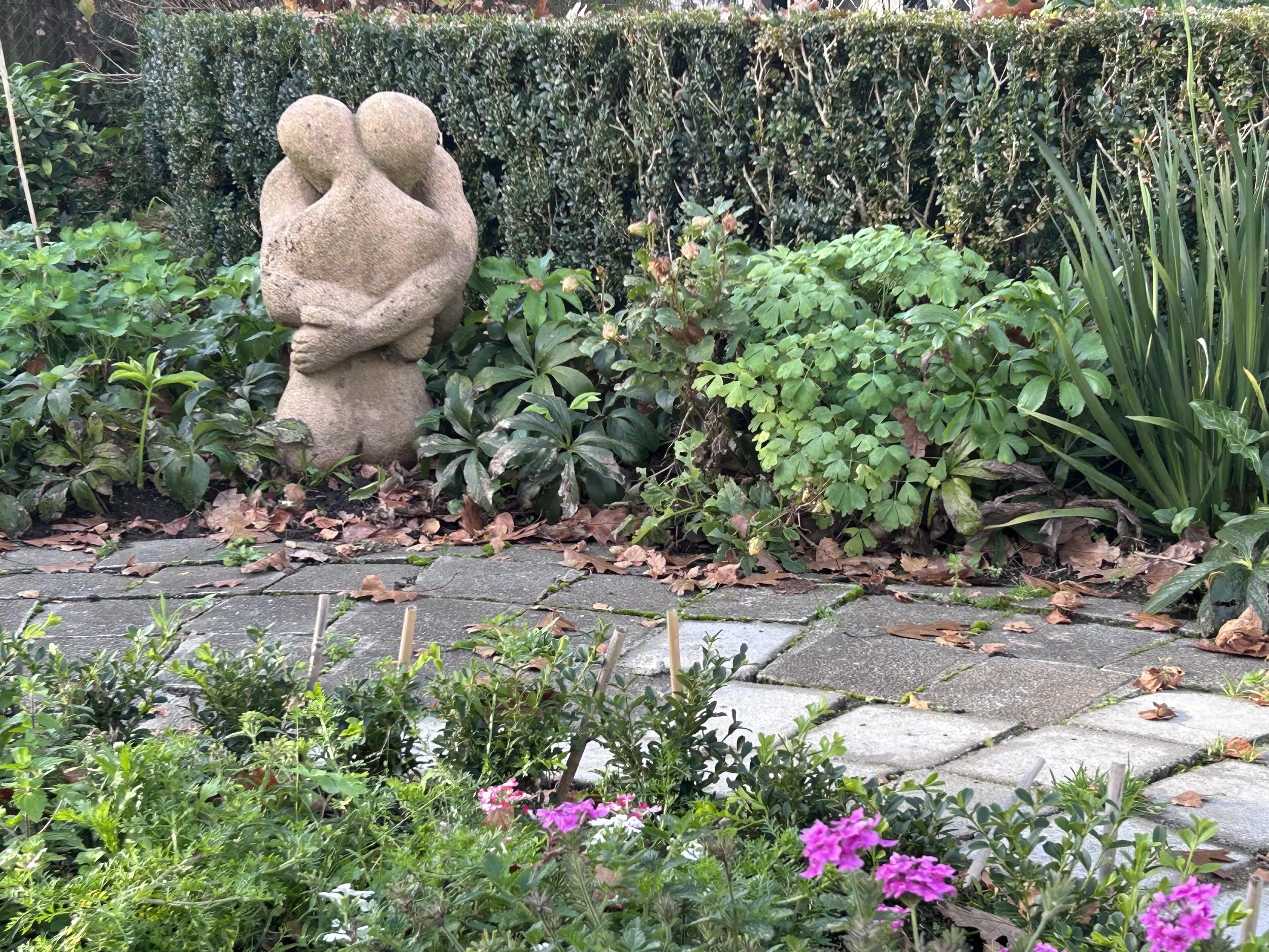 A concrete garden sculpture of two abstract, rounded figures embracing each other, surrounded by lush green bushes and plants with pink flowers and fallen leaves.