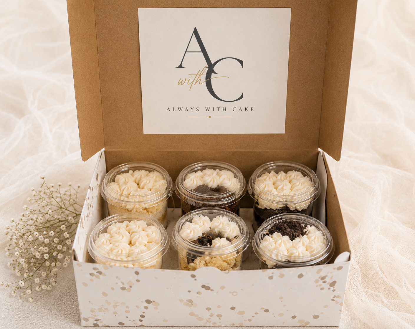 A box of six individual cupcakes with white frosting and cookie crumbles, some topped with Oreo cookies, on a light fabric surface with a sprig of baby's breath flower.