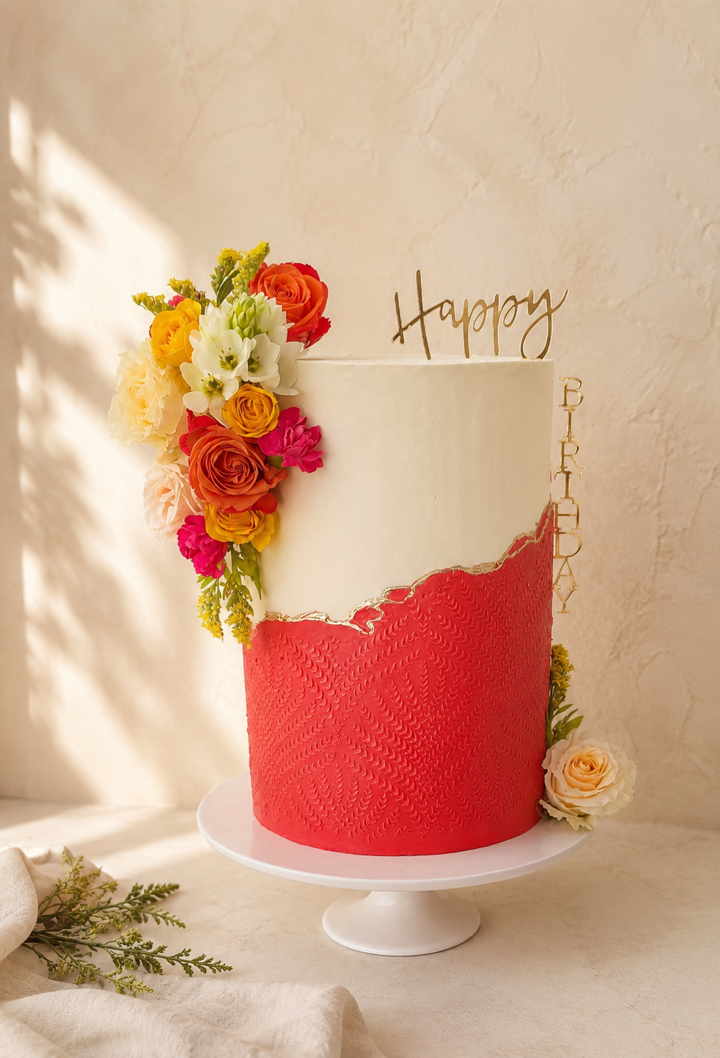 A tall birthday cake with white top and red bottom, decorated with colorful flowers on top and on the side, with a 'Happy Birthday' topper.