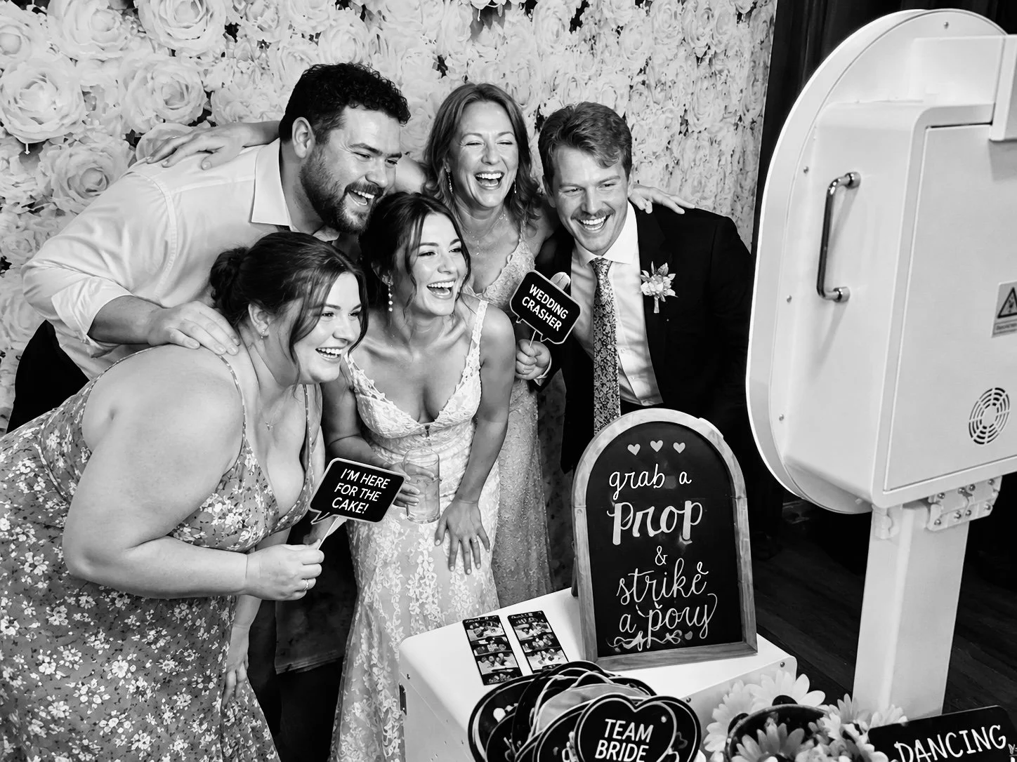 Group of people taking a photo in photo booth at a wedding, holding fun signs and smiling.