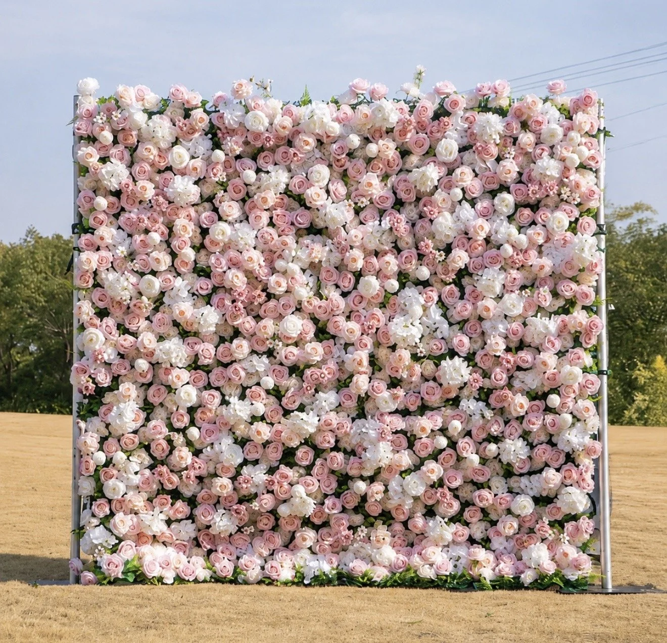 A large decorative floral wall made of pink and white artificial flowers outdoors with trees and a clear sky in the background.