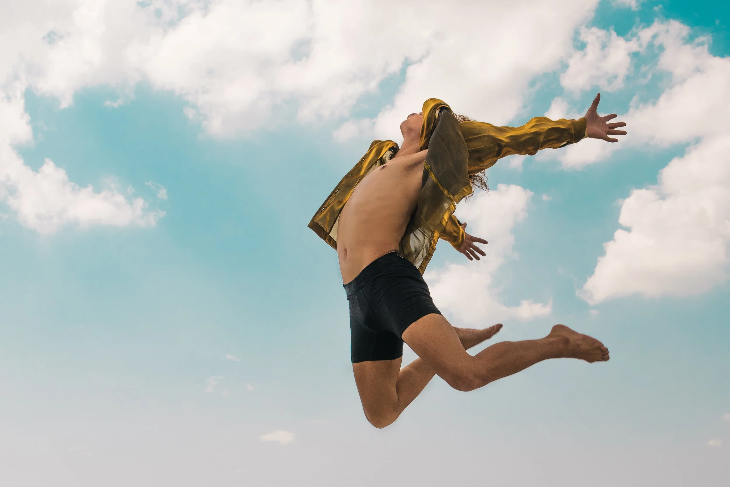 A person in shorts and a gold jacket is performing a leap against a partly cloudy sky, with arms and legs stretched outward.