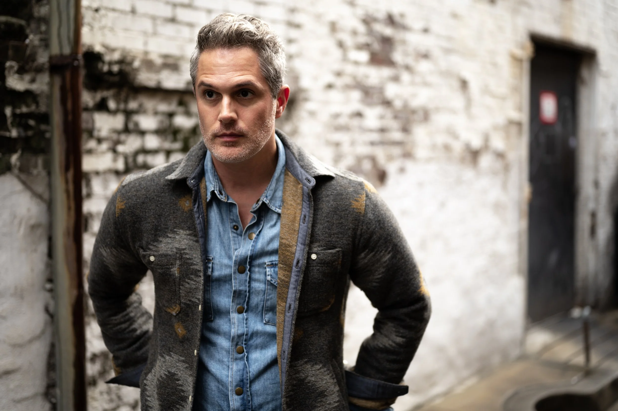 A man with gray hair and a short beard, wearing a denim shirt and a patterned cardigan, standing outdoors in front of a brick wall with out-of-focus doorway in the background.