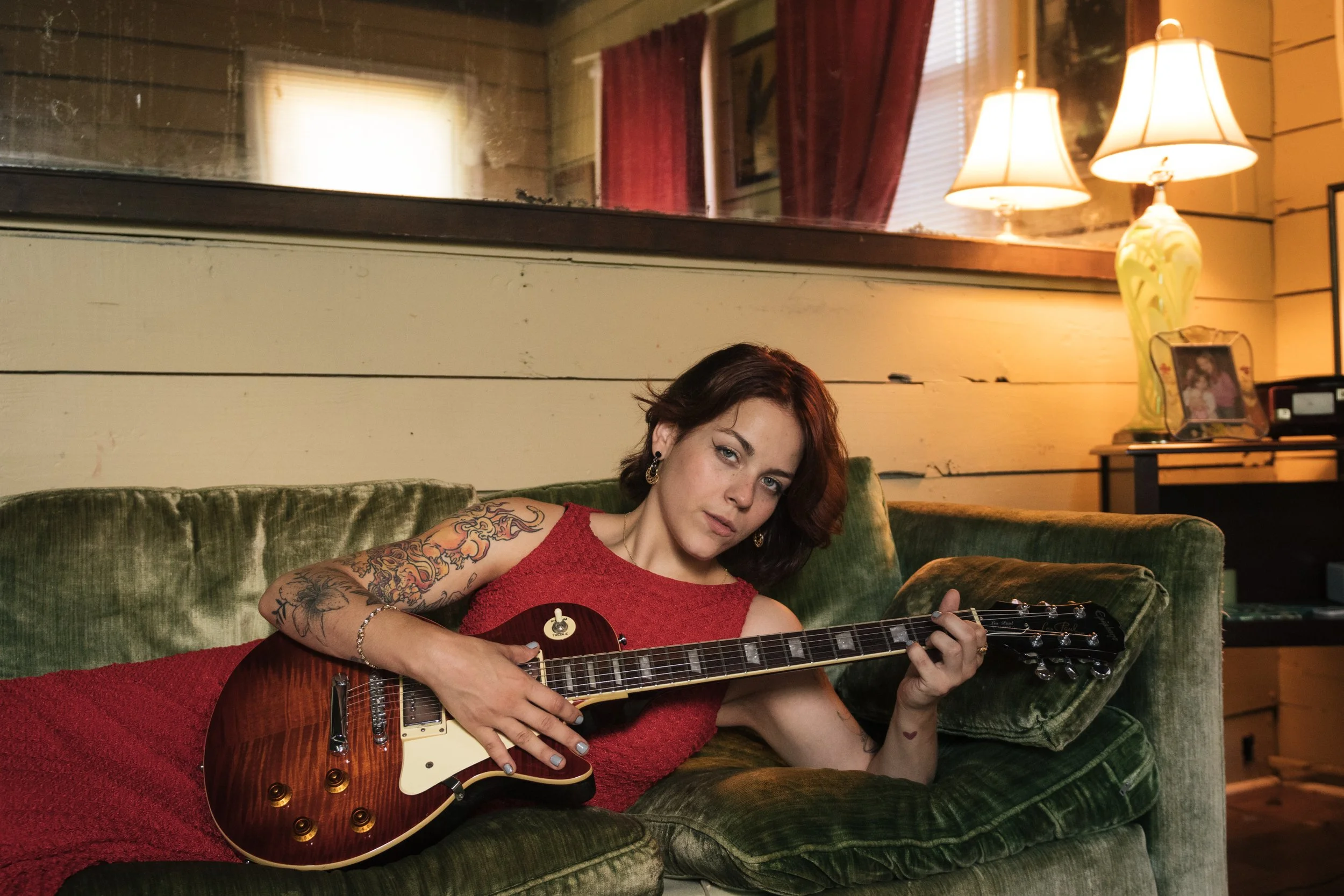 Young woman with tattoos and dark hair playing an electric guitar on a green velvet couch in a cozy, warmly lit living room.
