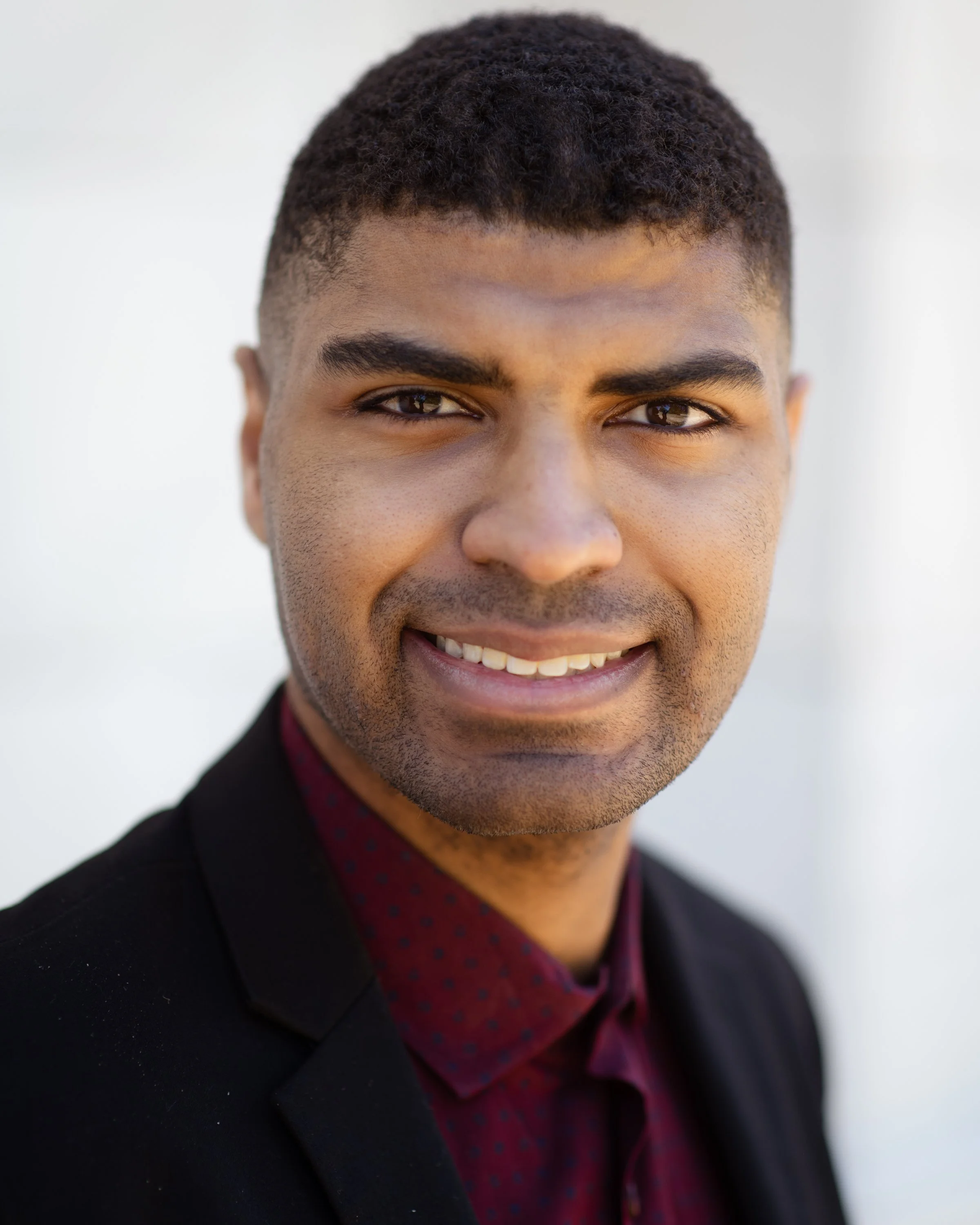 Close-up of a young man with short, curly black hair, smiling, wearing a dark blazer over a burgundy shirt with a subtle pattern, against a plain, light-colored background.