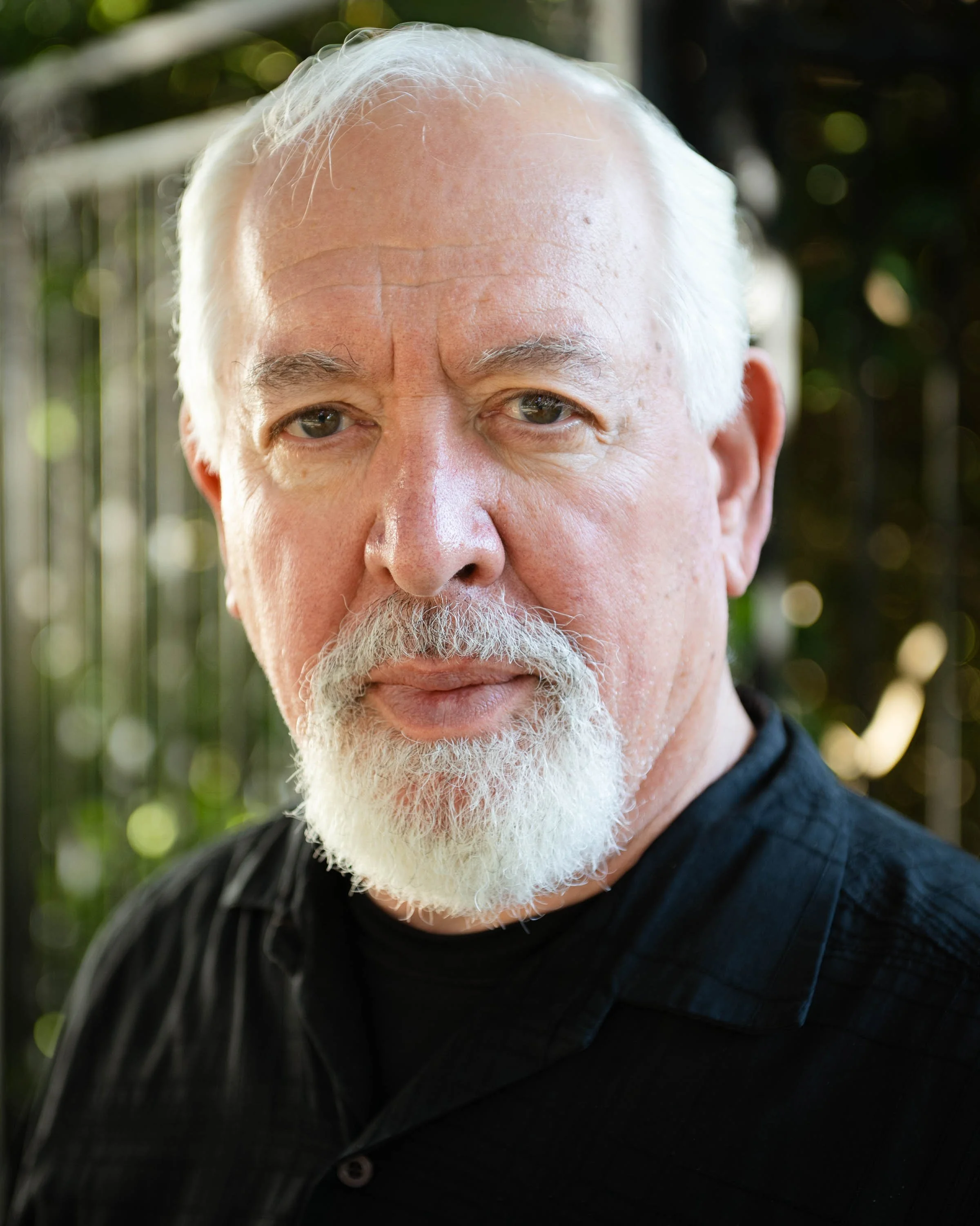Close-up of an elderly man's face, showing his gray hair, wrinkles, and beard, with a dark shirt and blurred green background.