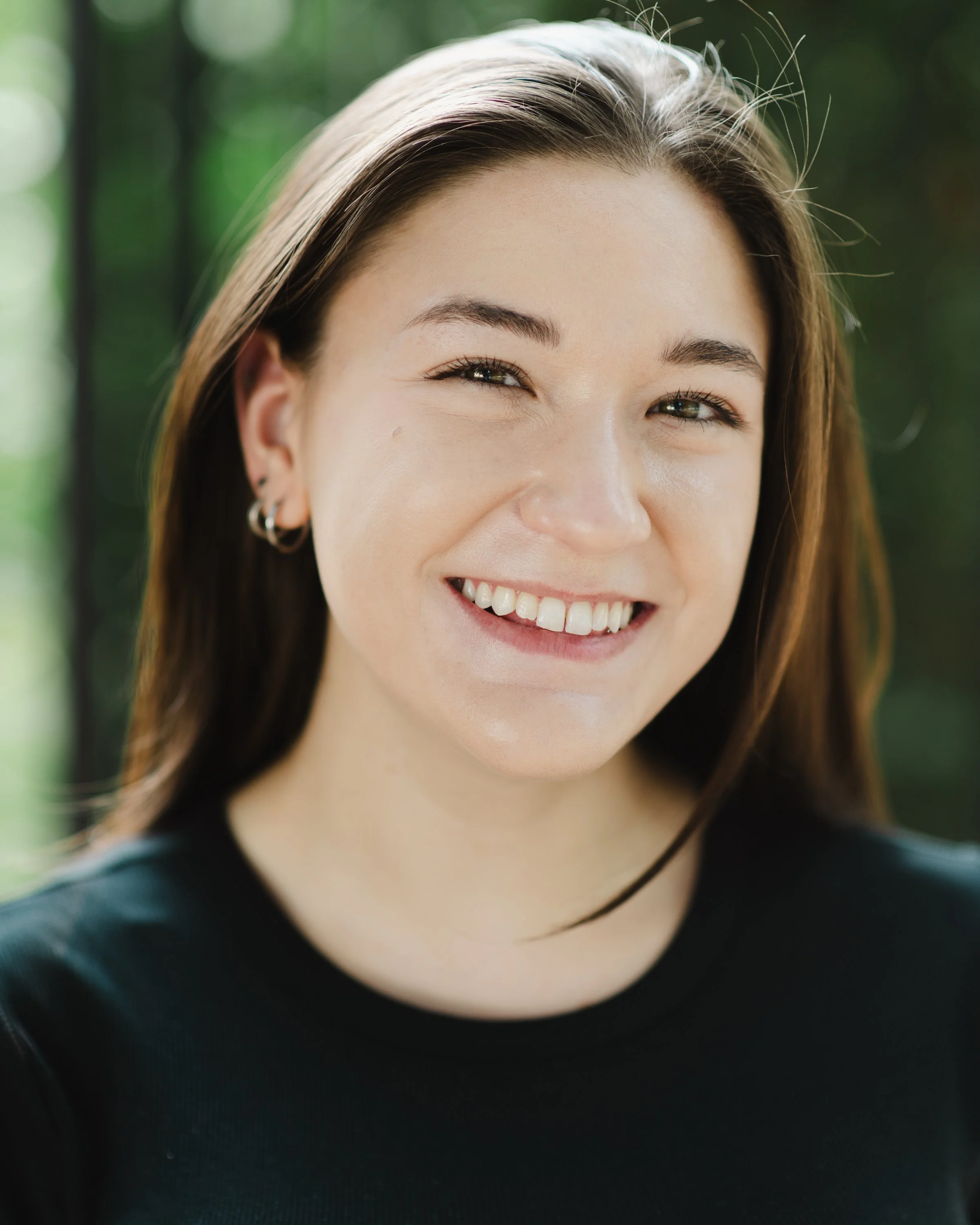 A smiling woman with long brown hair, hoop earrings, and a black shirt, outdoors with a blurred green background.