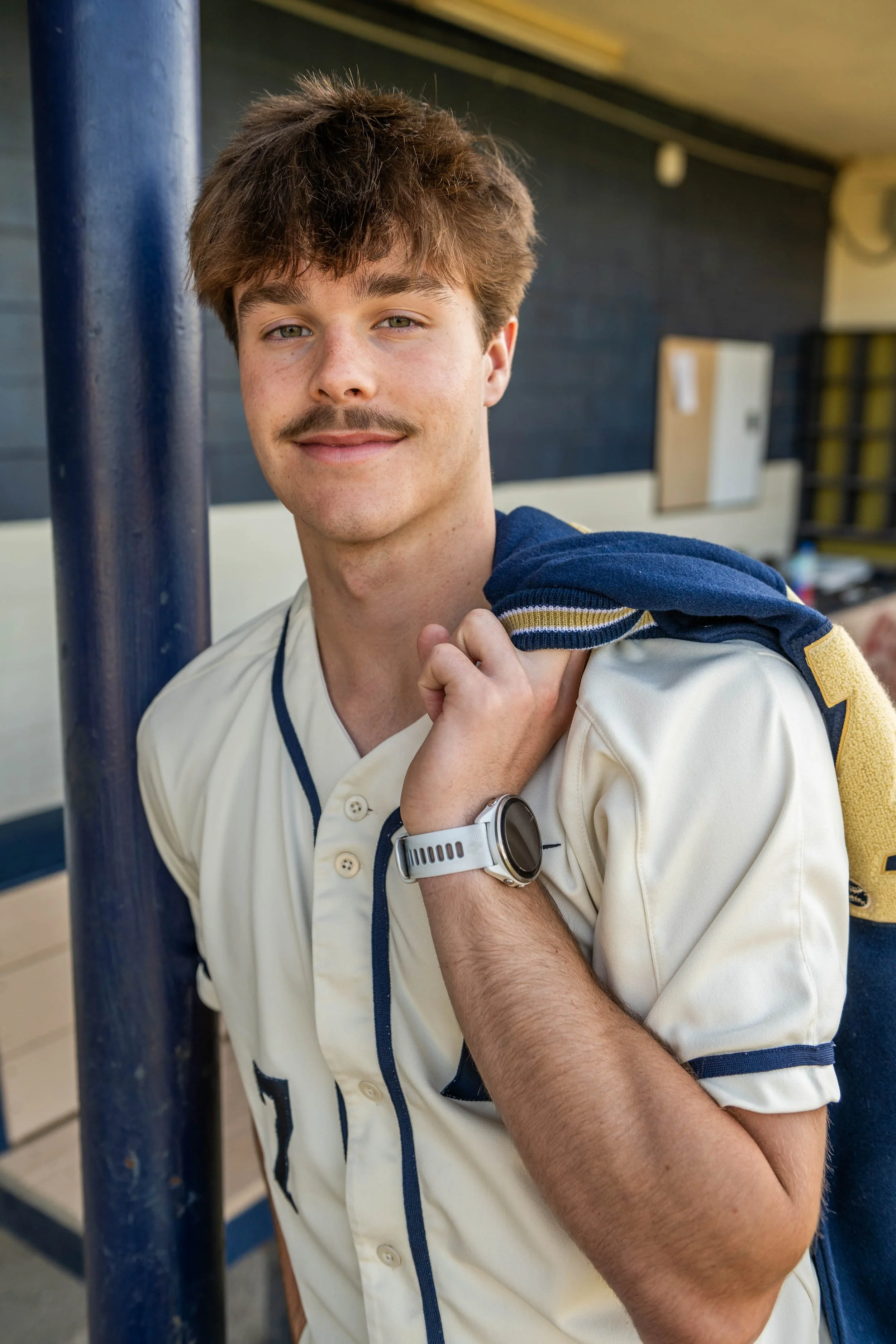 A young man with light brown hair, a mustache, and light skin, wearing a white sports jersey with navy blue accents and a white watch, holding a navy blue jacket over his shoulder, standing near a blue pole indoors.