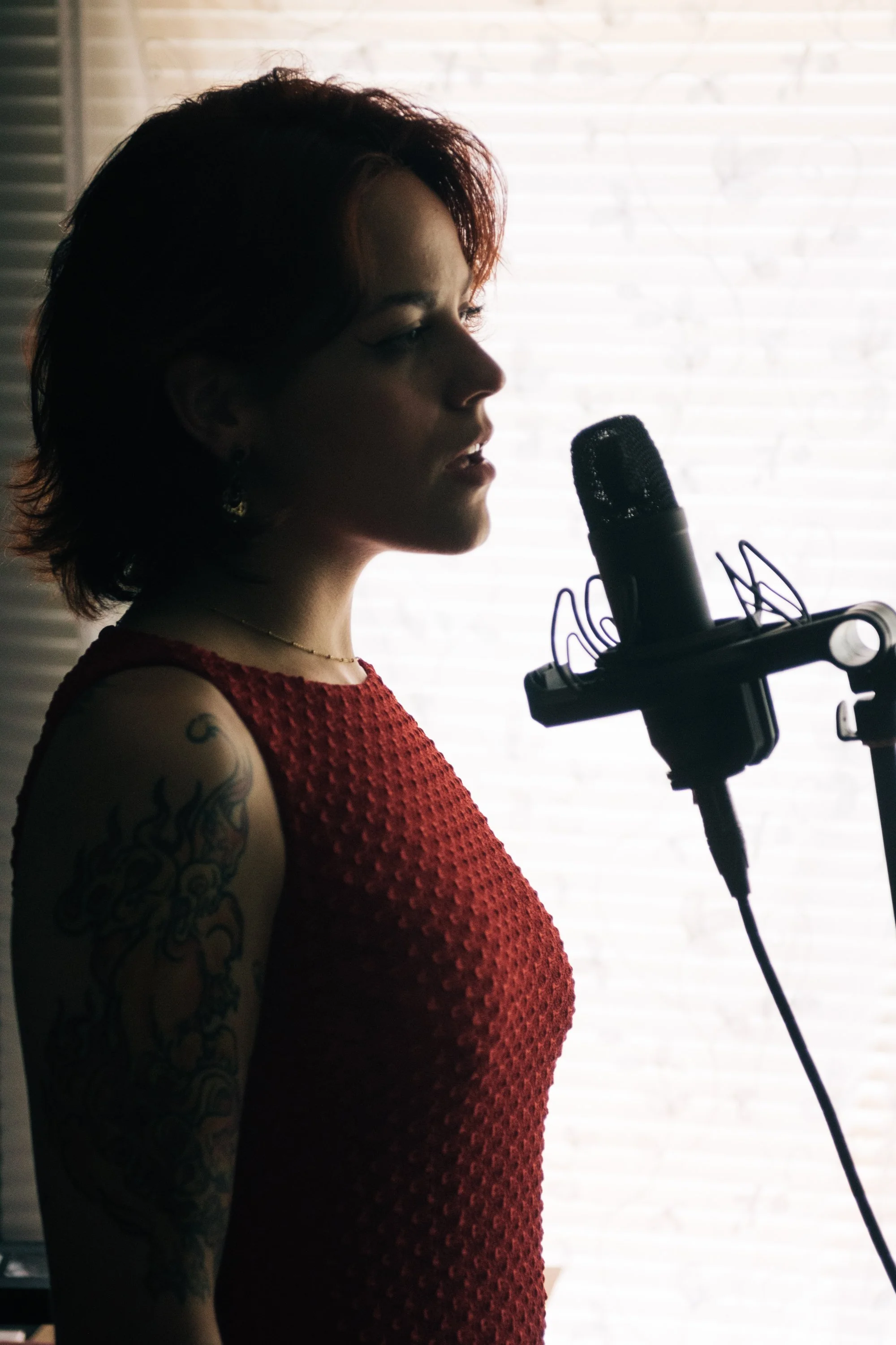 Woman with short dark hair and tattoos on her arm singing into a microphone in front of a backlit window.