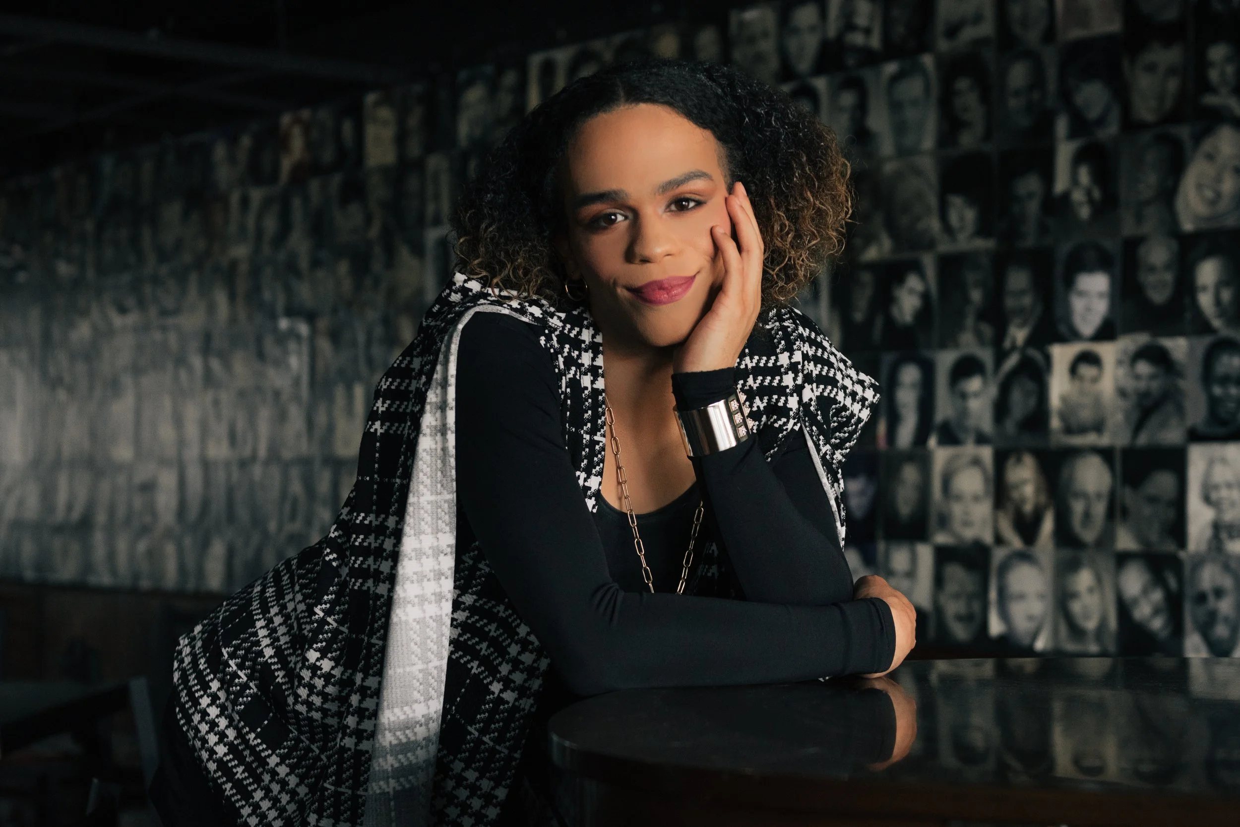 Portrait of a young person with curly hair, smiling gently, resting their face on their hand, seated at a table in front of a wall decorated with black and white photographs.