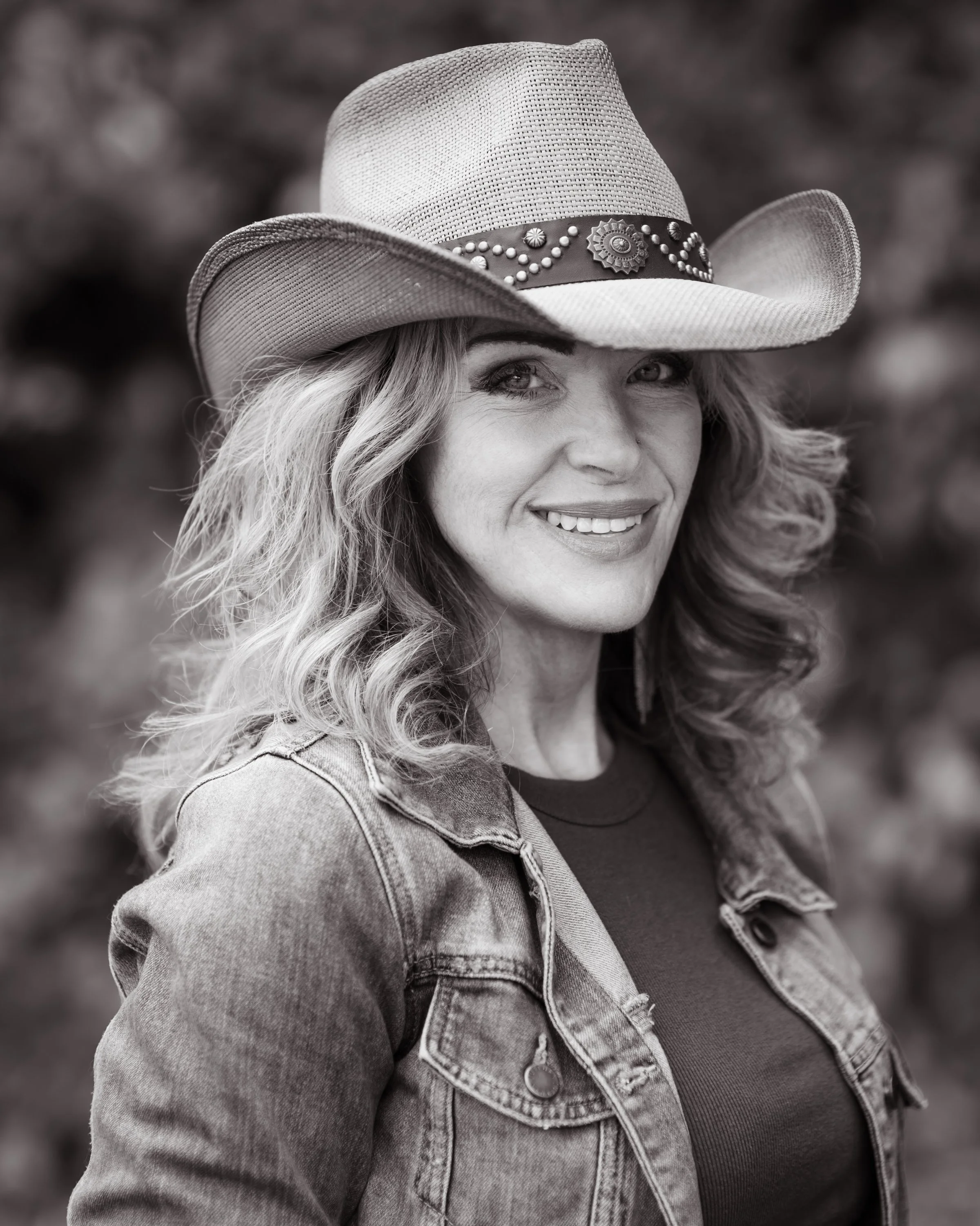Woman with wavy hair smiling wearing a cowboy hat with decorative band, denim jacket, and a dark shirt underneath.