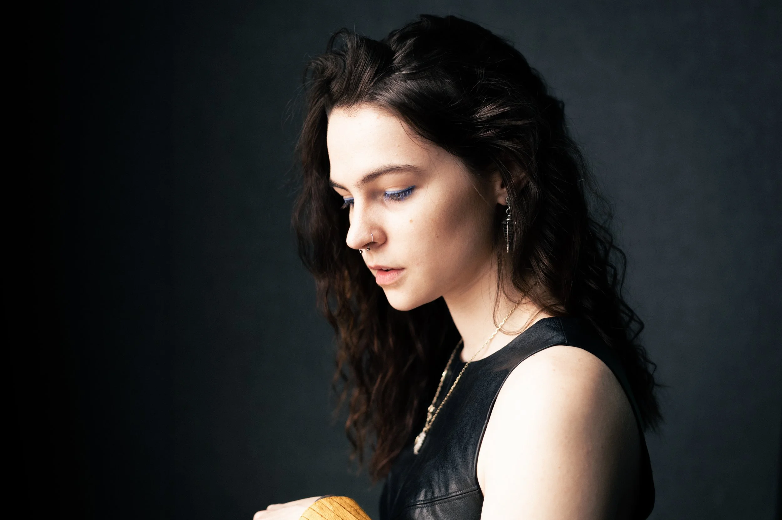 A young woman with dark, curly hair and light skin, wearing jewelry, looking down against a dark background.