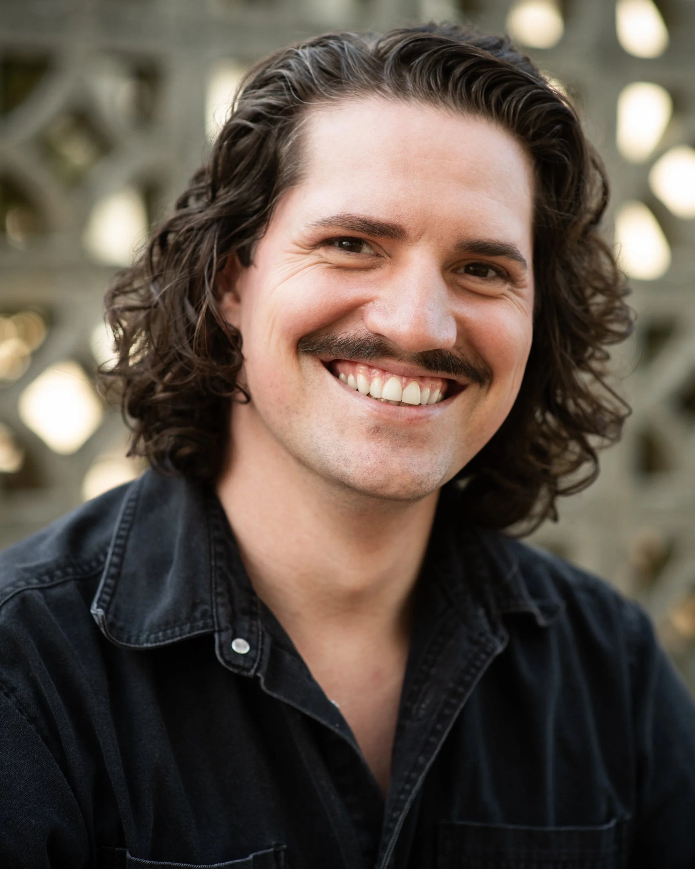 Close-up of a smiling person with dark curly hair and a mustache, wearing a black denim jacket.