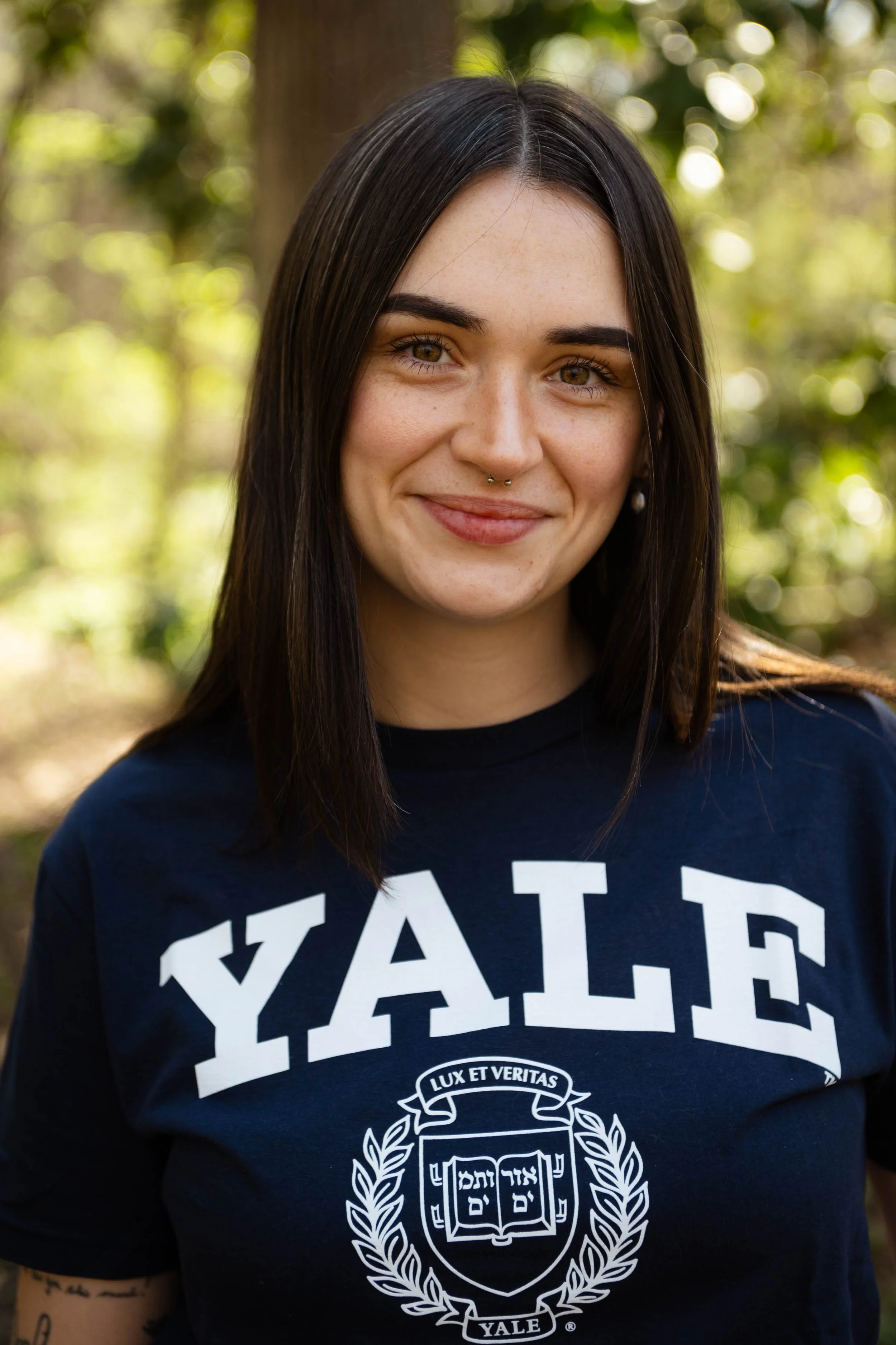 Close-up of a young woman with long dark hair, light skin, and a septum piercing, smiling outdoors with a blurred background of trees and sunlight, wearing a navy blue Yale sweatshirt.
