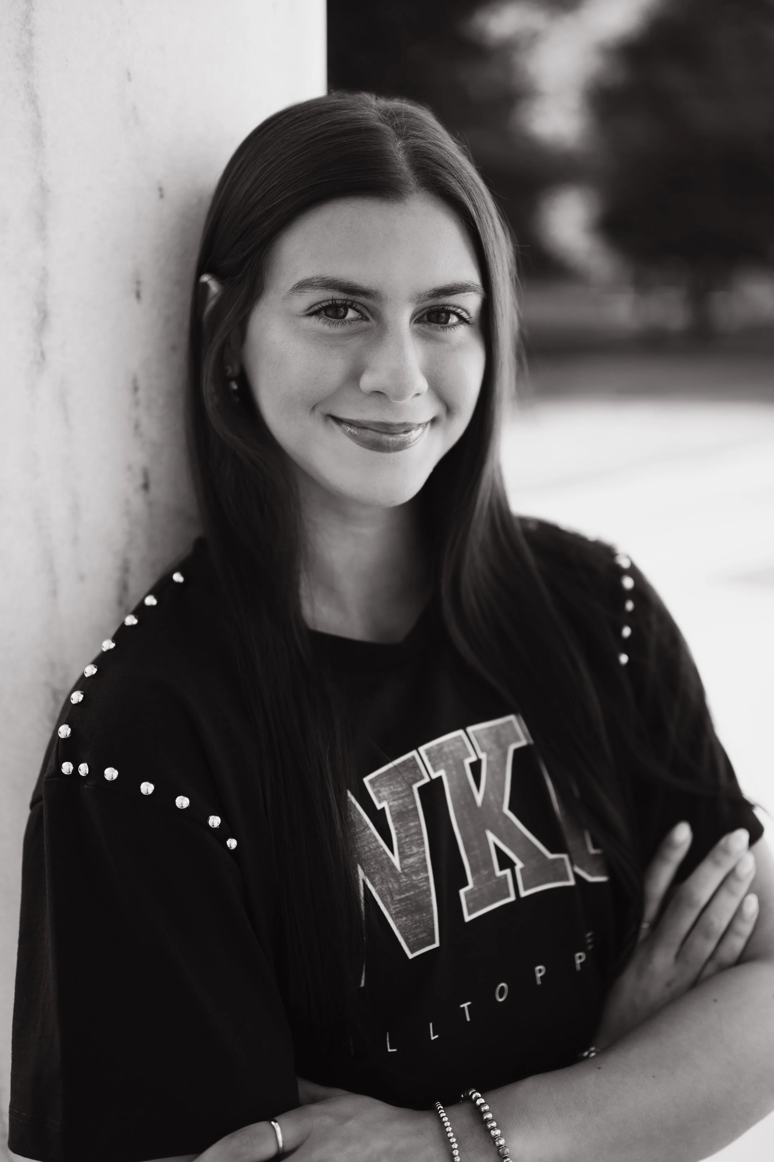 A young woman with long dark hair, smiling softly, leaning against a wall outdoors, wearing a black shirt with metal studs on the shoulders and a printed graphic, with her arms crossed and jewelry on her wrists.
