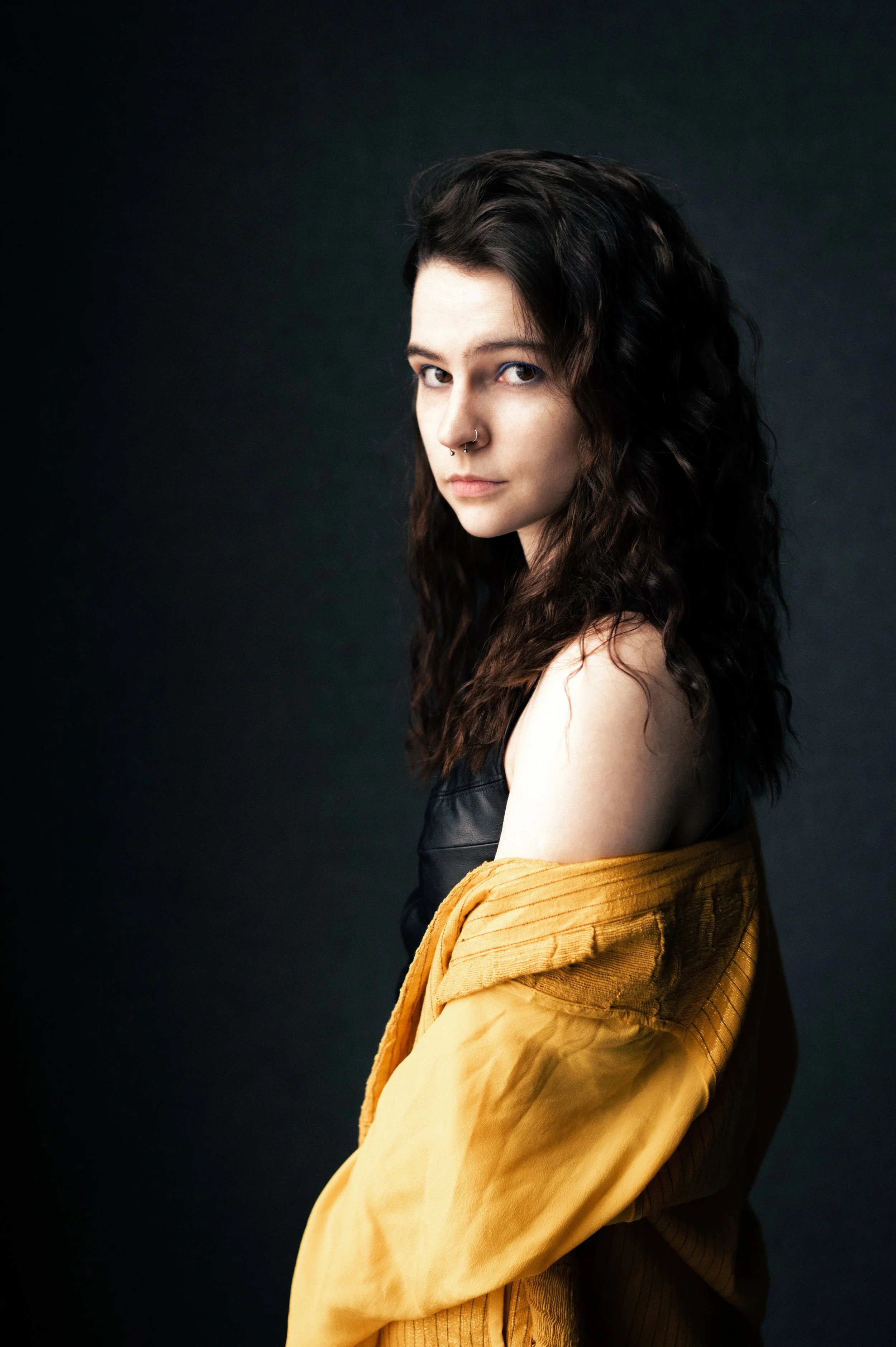 A young woman with wavy dark hair, wearing a black top and a mustard-yellow jacket, looks confidently at the camera against a plain dark background.