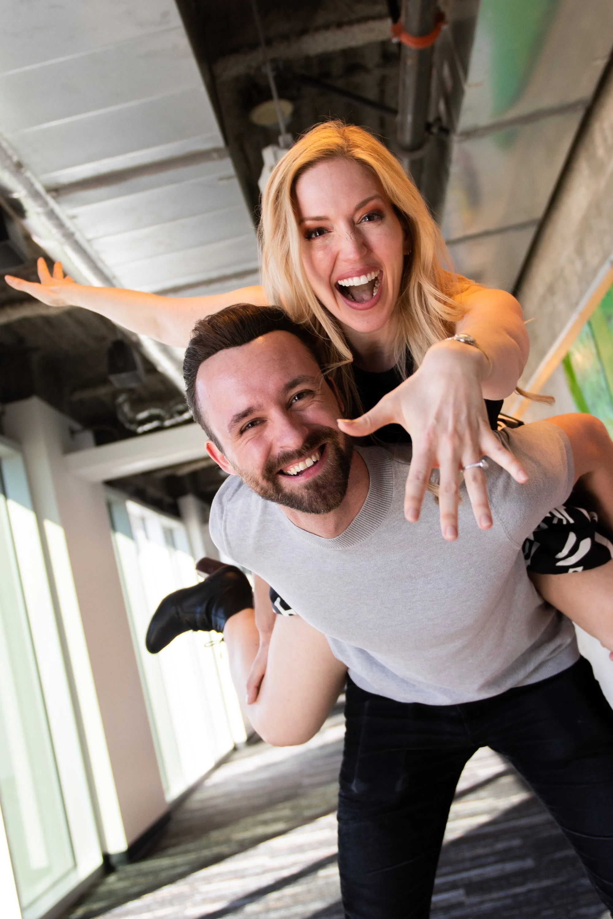 A woman with blonde hair and a man with a beard are playing and smiling inside a modern building with large windows, hardwood floors, and an industrial ceiling.