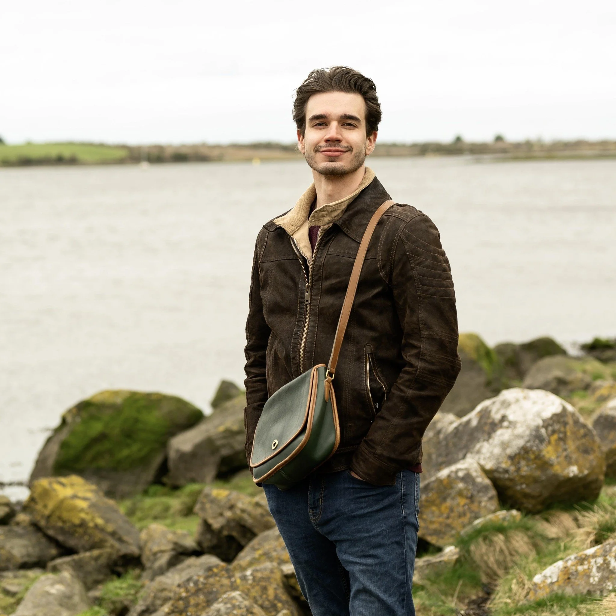 A young man with dark hair and a slight beard stands outdoors near a body of water with rocks and greenery in the background, smiling at the camera.
