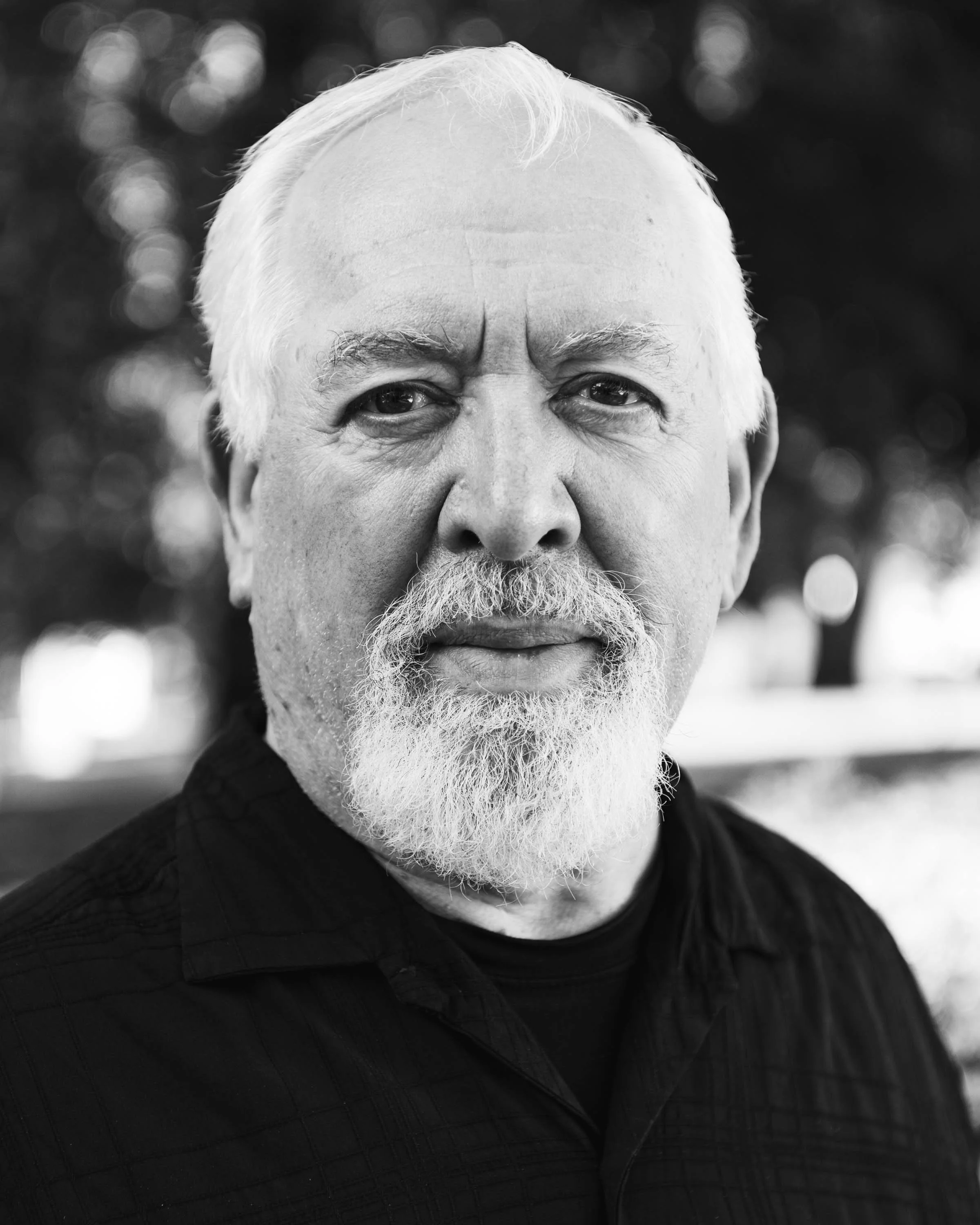 Close-up of an elderly man with white hair and beard, wearing a dark quilted jacket, outdoors with blurred background of trees or foliage.